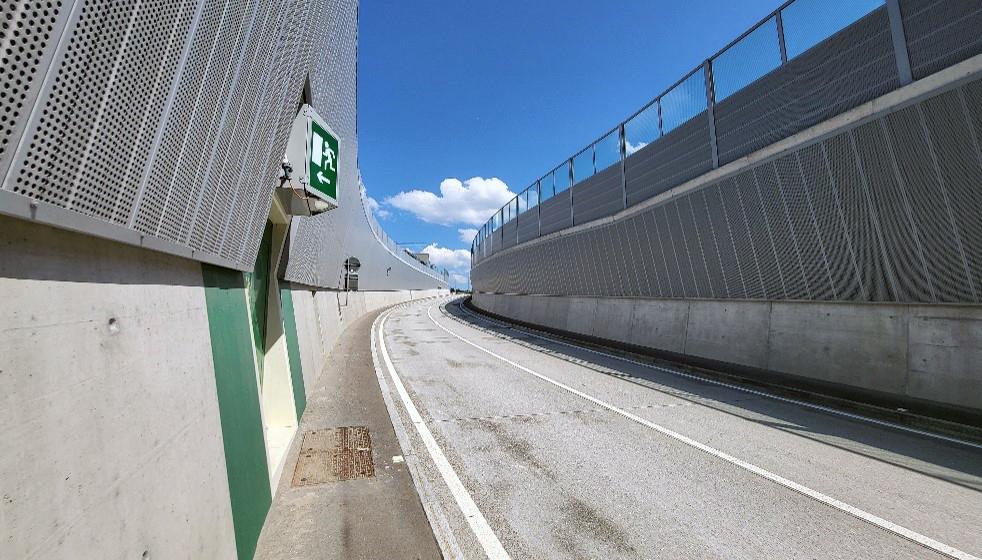 Gekrümmte Strasse mit hohen Metallgitterbarrieren auf beiden Seiten. Der asphaltierte Weg ist mit weissen Linien markiert und führt in eine Kurve und leicht bergauf. Links ist ein grünes Notausgangsschild zu sehen, das in Richtung des Weges zeigt.