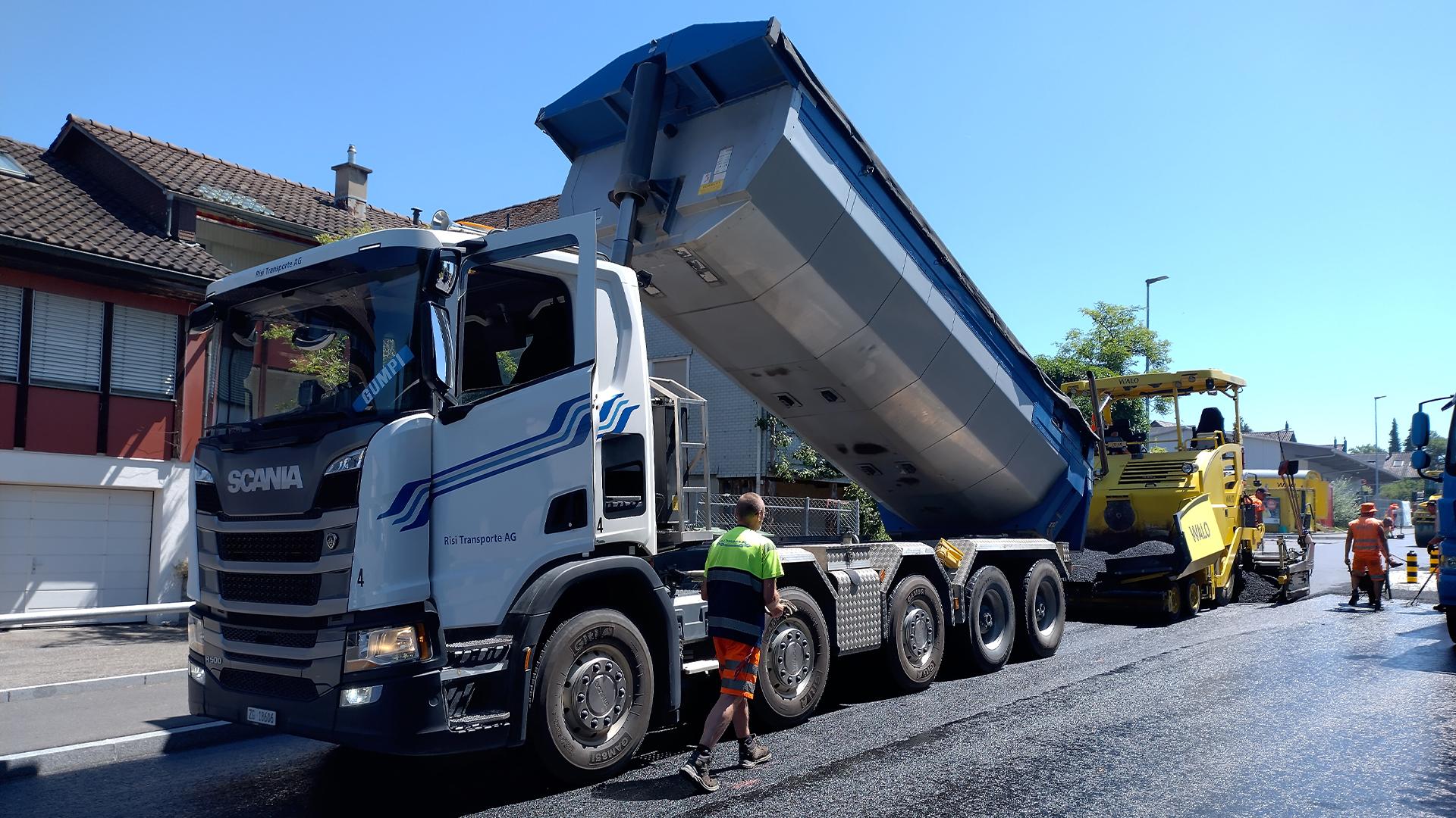 Strassenbaustelle mit weiss-blauem Kipper, der Asphalt in eine gelbe Strassenfertigungsmaschine kippt. Arbeiter in Warnkleidung, frischer Belag und Wohnhäuser im Hintergrund bei sonnigem Wetter.