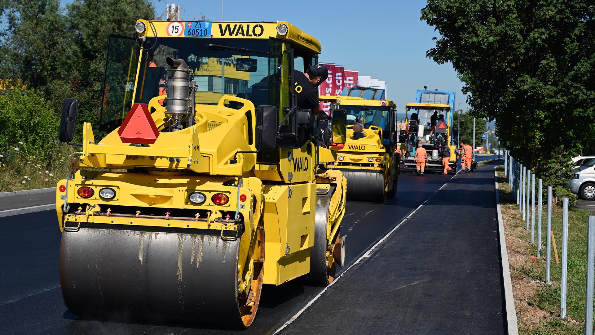 Strassenbaustelle mit mehreren gelben Walzen der Firma WALO und Arbeitern in orangefarbener Kleidung beim Asphaltieren einer neuen Strasse.
