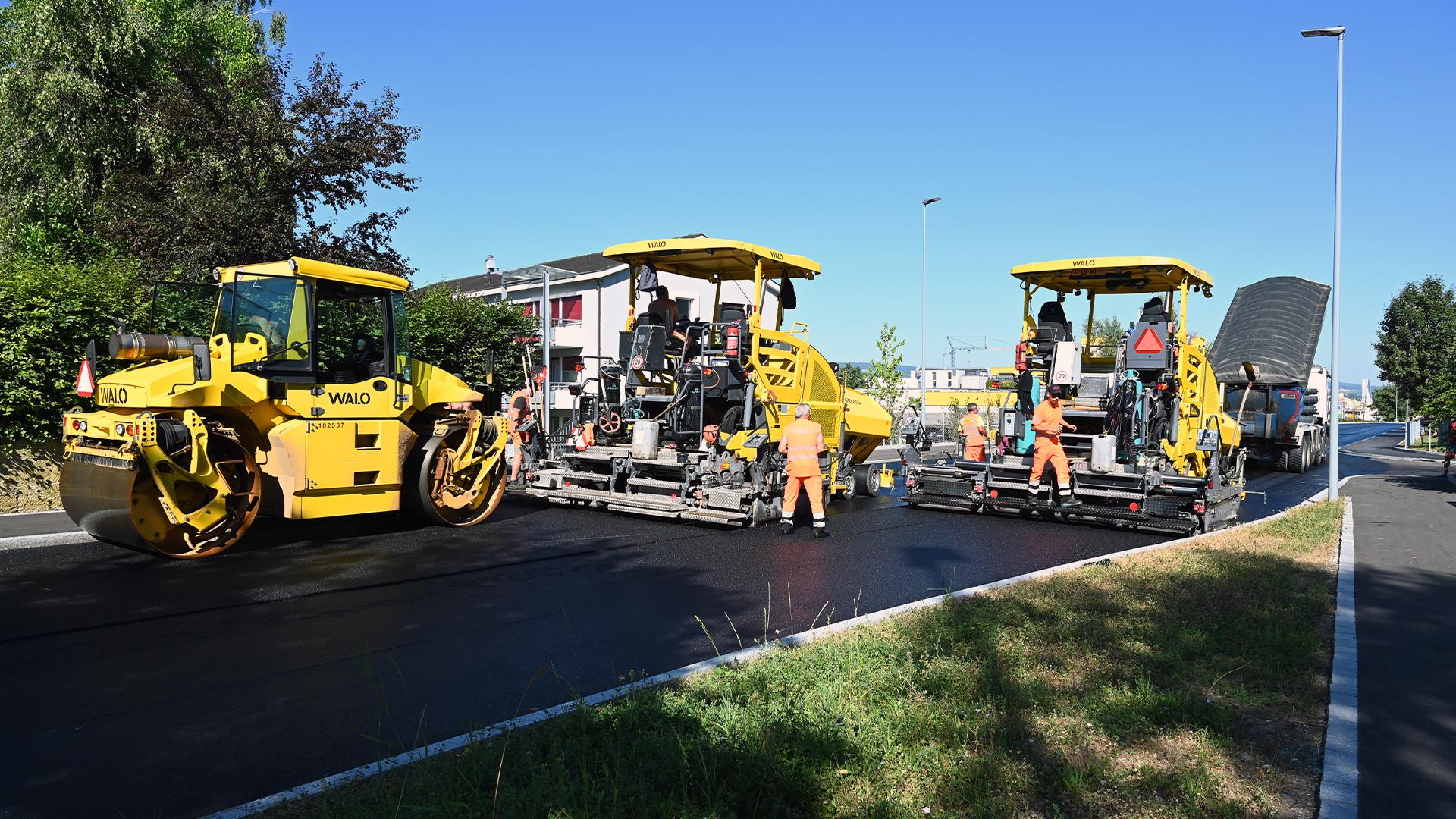 Strassenbaustelle mit gelben Asphaltfertigern und einer Walze der Firma WALO, betrieben von Arbeitern in orangefarbener Kleidung. Ein Lastwagen kippt Material in einen Fertiger.