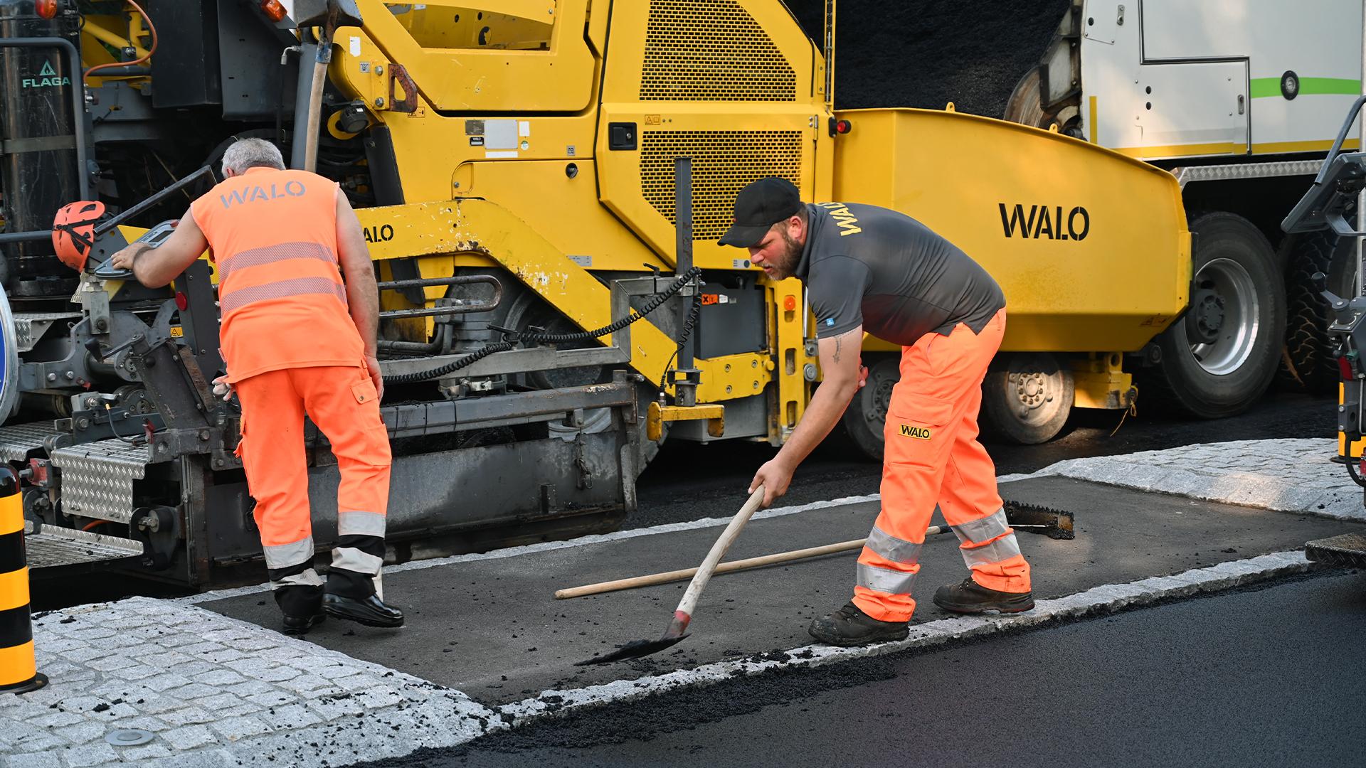 Zwei WALO-Arbeiter in orangefarbener Kleidung verteilen Asphalt auf einer Baustelle – einer mit einer Maschine, der andere mit einer Schaufel. Im Hintergrund steht ein Lastwagen mit Asphalt.