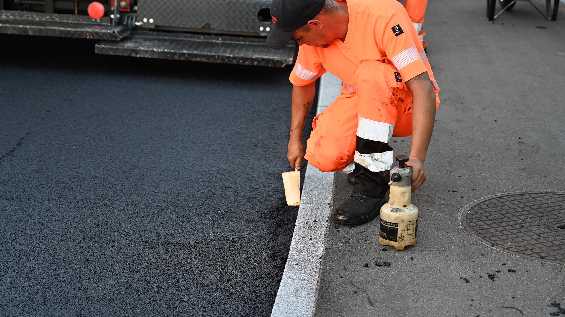 Ein Strassenarbeiter in orangefarbener Kleidung glättet mit einem Handwerkzeug den Rand des frisch asphaltierten Belags entlang des Randsteins.