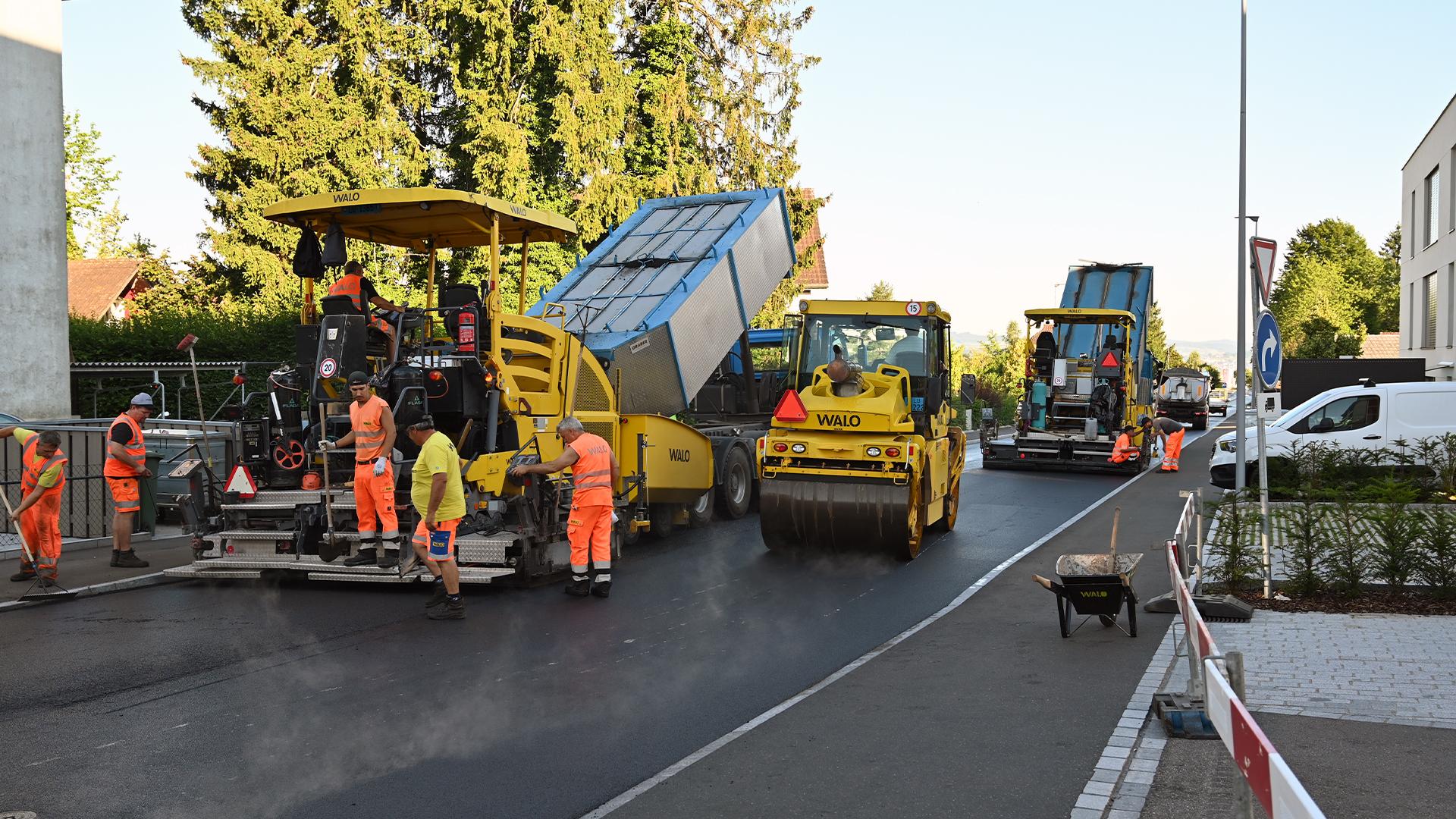 Mehrere Arbeiter in orangefarbener Kleidung arbeiten mit Walzen, Asphaltfertiger und weiteren Maschinen an einer Strassenbaustelle.