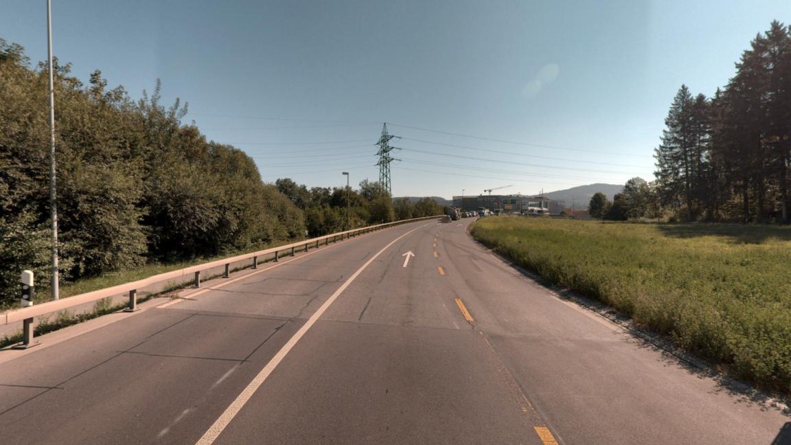 Strassenabschnitt mit Fahrbahnmarkierungen und Leitplanken, umgeben von Wiesen und Bäumen.