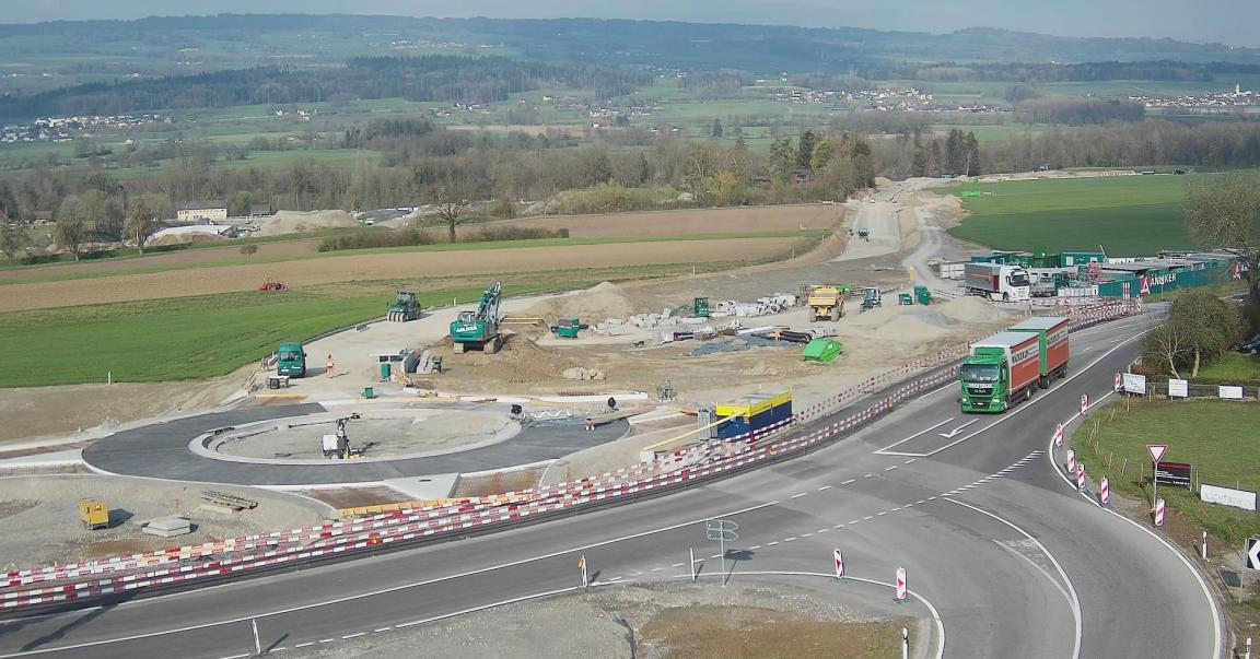 Das Bild zeigt eine Strassenbaustelle in ländlicher Umgebung. Der Kreisverkehr befindet sich im Bau. Lastwagen und Baumaschinen sind auf der Baustelle sichtbar. Im Hintergrund erstreckt sich eine hügelige Landschaft mit Feldern und Wäldern.