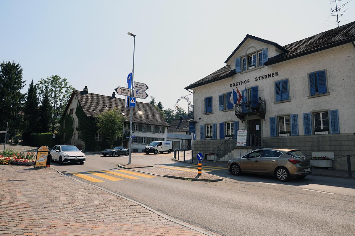 Blick auf die eine Strassenkreuzung mit einem angeschriebenen Gebäude rechts sowie einem Kandelaber mit Wegweisern sowie Autos und gelben Fussgängerstreifen.
