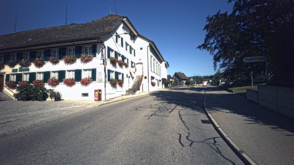 Aufwärts führende Strasse mit einem Haus auf der linken Seite und rechts einem Trottoir und einem Laubbaum.