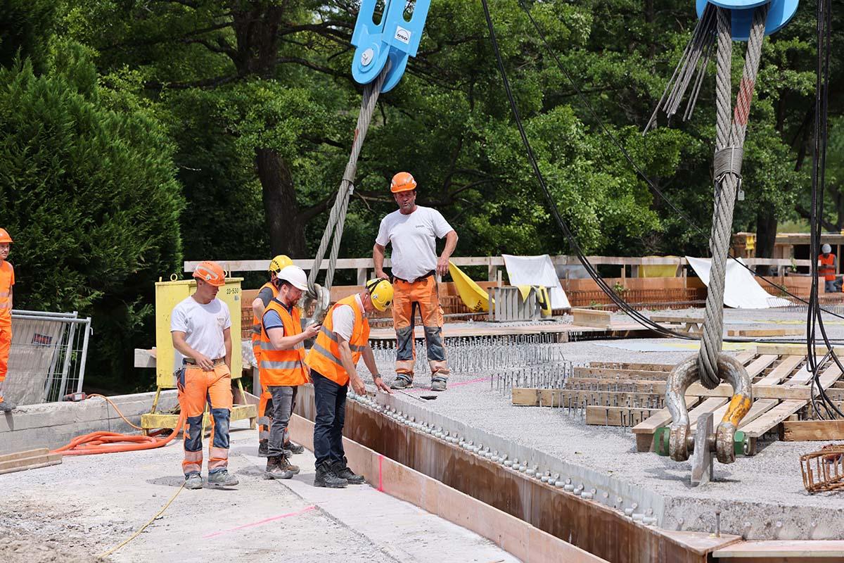 Bauleute in Leuchtwesten beobachten eine unfertige Betonbrücke, die an zwei Seilen hängt.