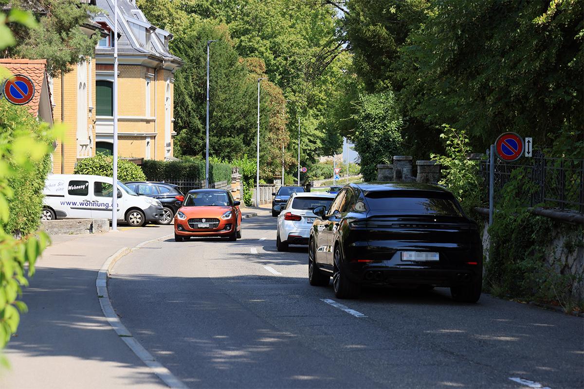 Mehrere Autos, darunter ein oranger Kleinwagen und ein schwarzer SUV, fahren hintereinander durch eine schattige Strasse mit Altbau-Villen.