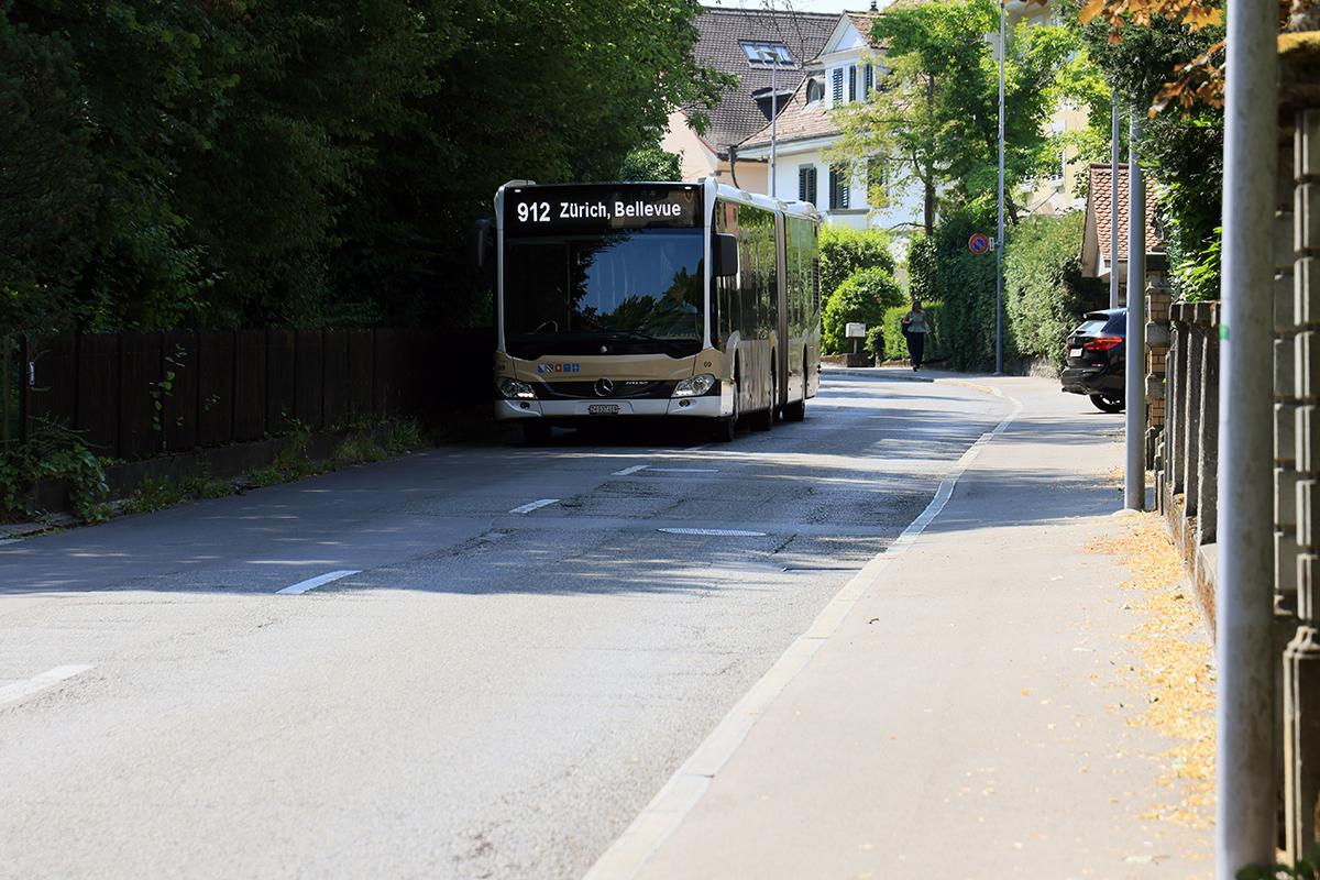 Ein beigefarbener Gelenkbus der Linie 912 fährt auf einer asphaltierten Strasse an Wohnhäusern und Gehwegen vorbei.