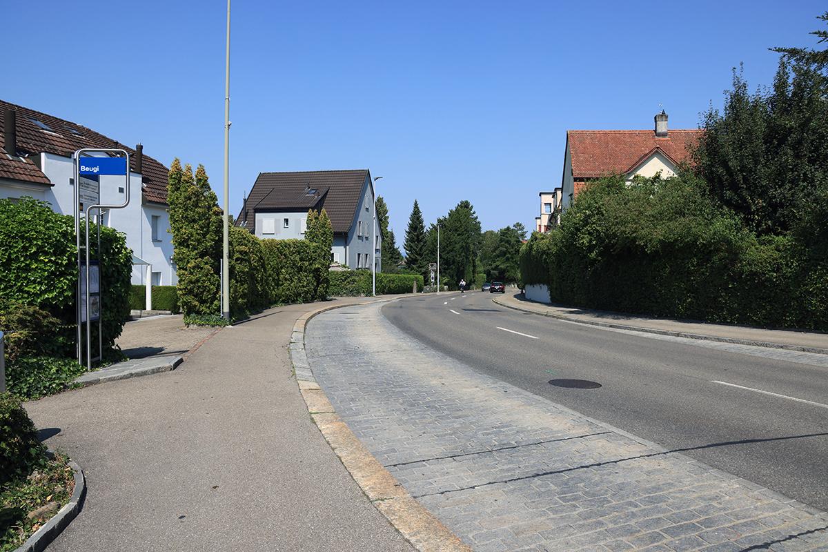 Das blaue Haltestellenschild «Beugi» an einem gepflasterten Strassenabschnitt, gesäumt von Wohnhäusern und gepflegten Hecken.