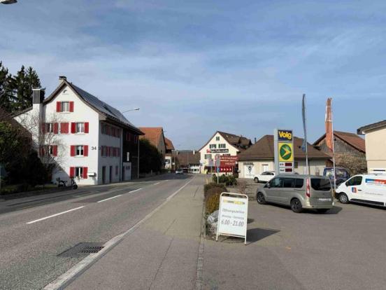 Ein Abschnitt der Zürcherstrasse in Lufingen. Links der Strasse ein helles Wohnhaus mit roten Fensterläden, rechts ein Parkplatz und ein Tankstellen- und Volg-Schild. Im Hintergrund weitere Häuser des Dorfes.
