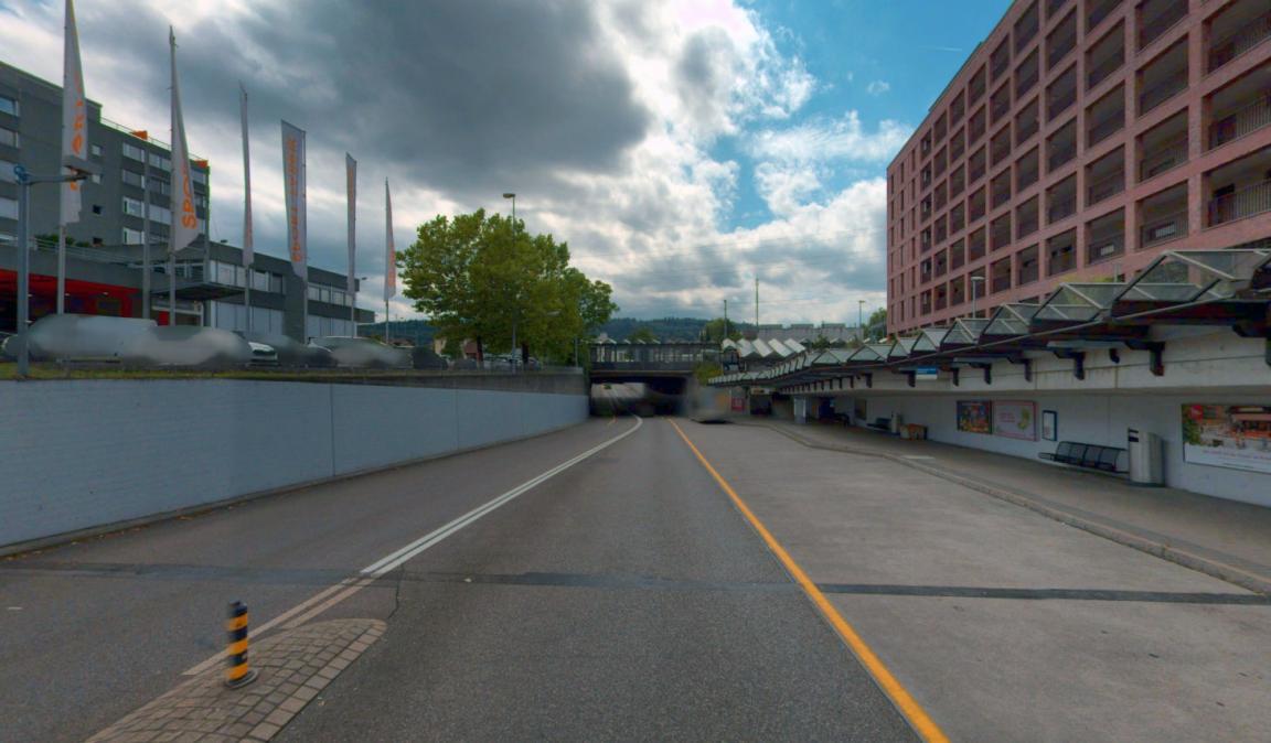 Eine zweispurige Strasse führt unter einer Eisenbahnbrücke hindurch. Rechts befindet sich ein moderner, überdachter Busstopp vor einem grossen, rötlichen Wohn- und Geschäftshaus. Links sind Werbefahnen und ein graues Bürogebäude zu sehen. Der Himmel ist bewölkt.
