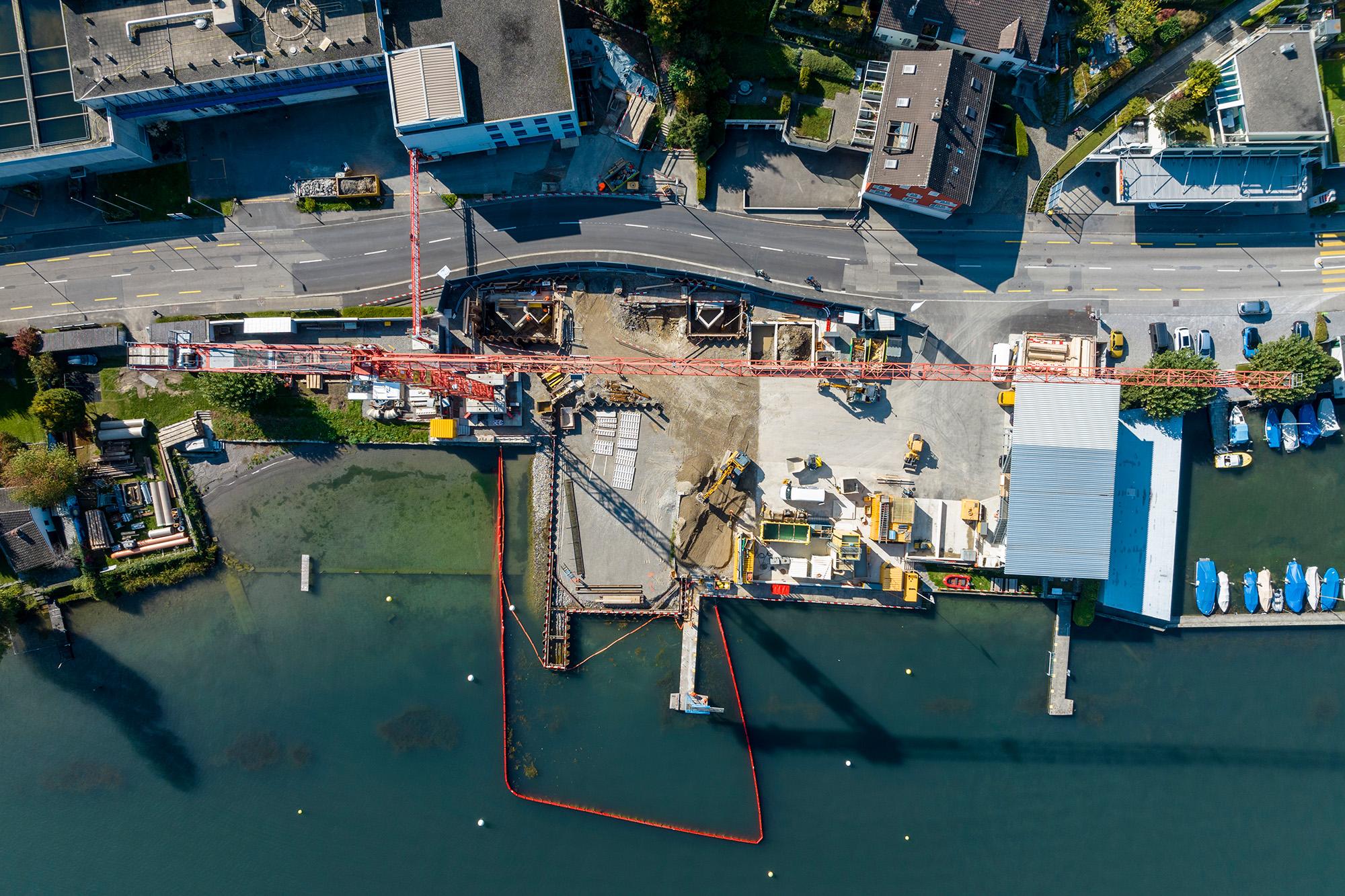 Vogelperspektive auf die Baustelle am Zürichsee. Vorne der See, in der Mitte die Baustelle mit  mehreren Baumaschinen und zwei Kranen, eine Strasse und im oberen Drittel Wohnhäuser.