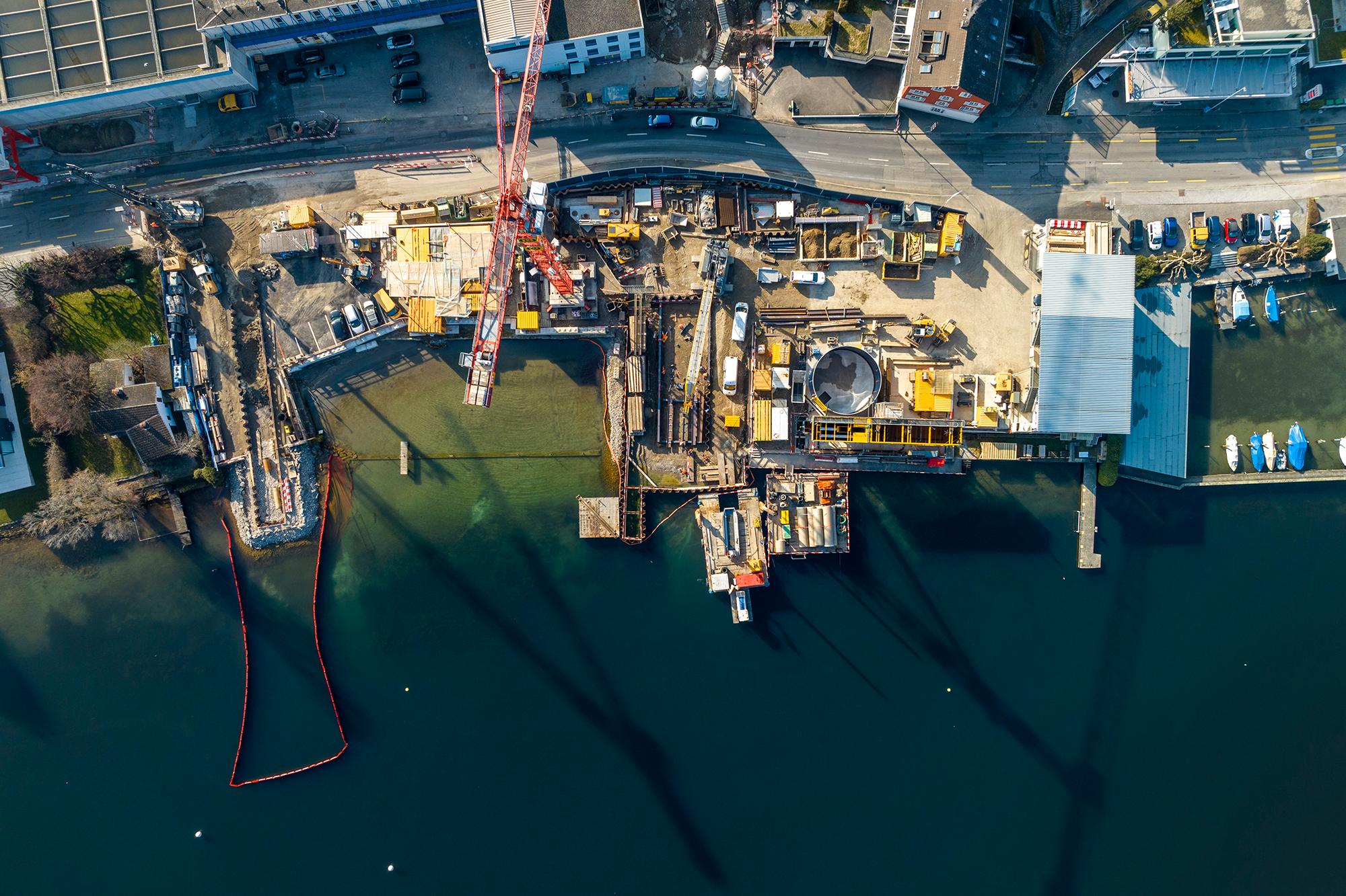Luftaufnahme des Zürichseeufers in Thalwil. Im unteren Teil des Bildes ist der See zu sehen, im oberen Drittel die Baustelle und dahinter die Strasse. Am prominentesten stehen ein roter Kran, die Baugrube und ein grosser Waschtank hervor