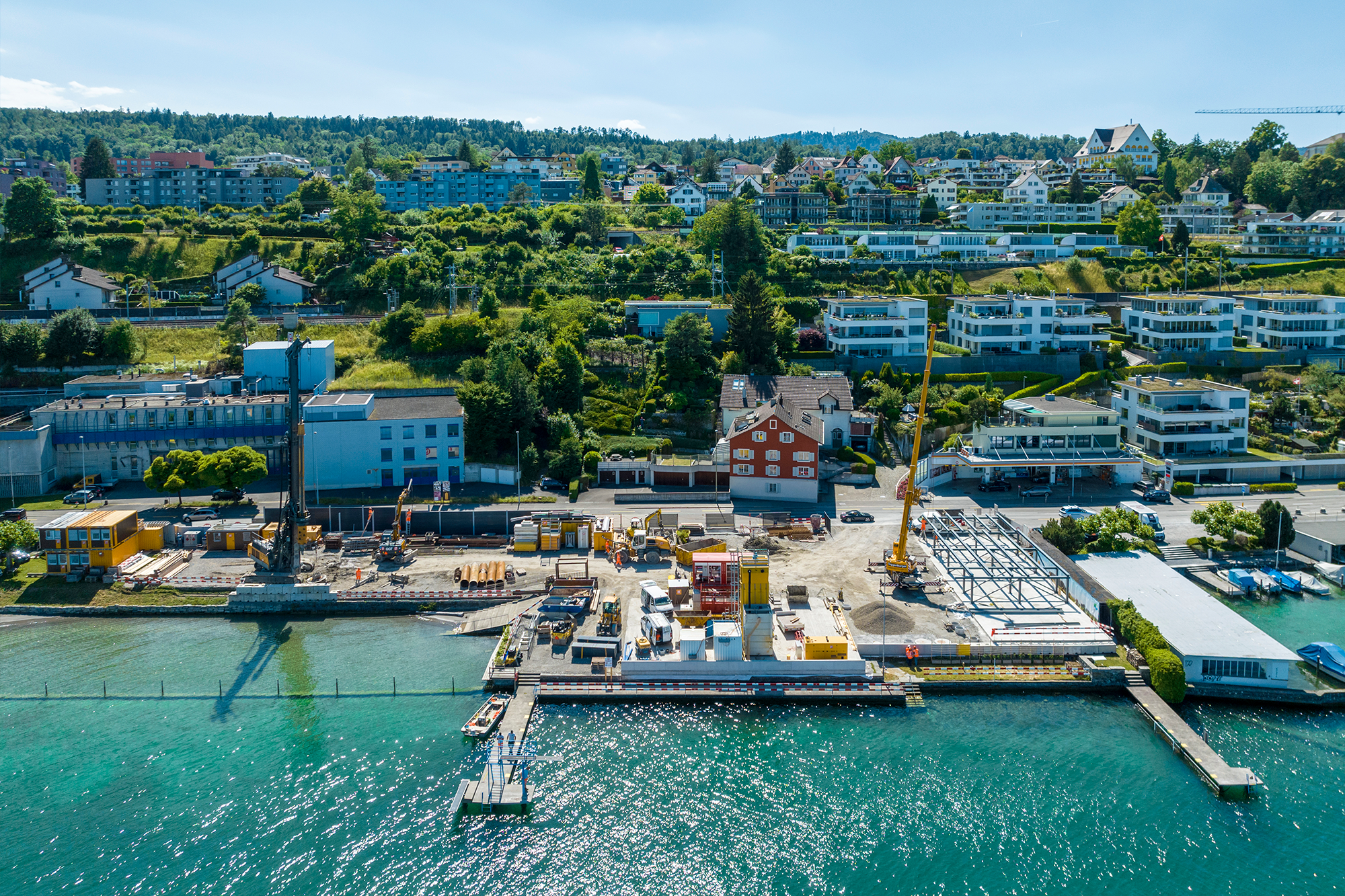 Vorne der türkise See, in der Mitte die Baustelle mit mehreren Baumaschinen und Einrichtungen, dahinter ein Hang mit mehreren Wohnhäusern. Blauer Himmel, die Sonne scheint.