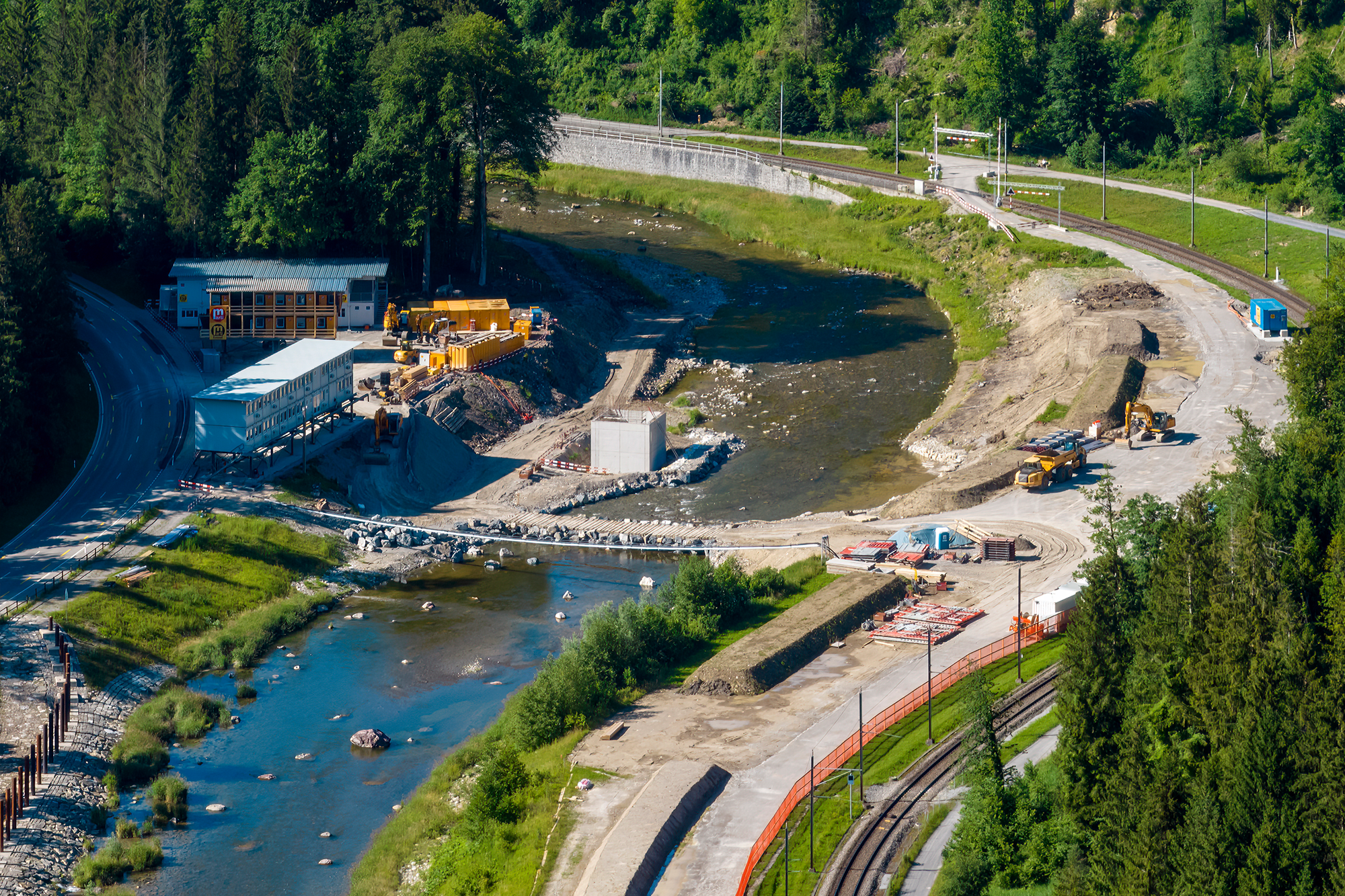 In der Mitte die Sihl, links die Sihltalstrasse, rechts die Bahnlinie der SZU. An der Strasse stehen Bürocontainer, durch die Sihl führt eine Furt, über die Baufahrzeuge den Fluss überqueren können. Zwischen Fluss und Bahn sind grosse Flächen für Bauinstallationen freigeräumt.