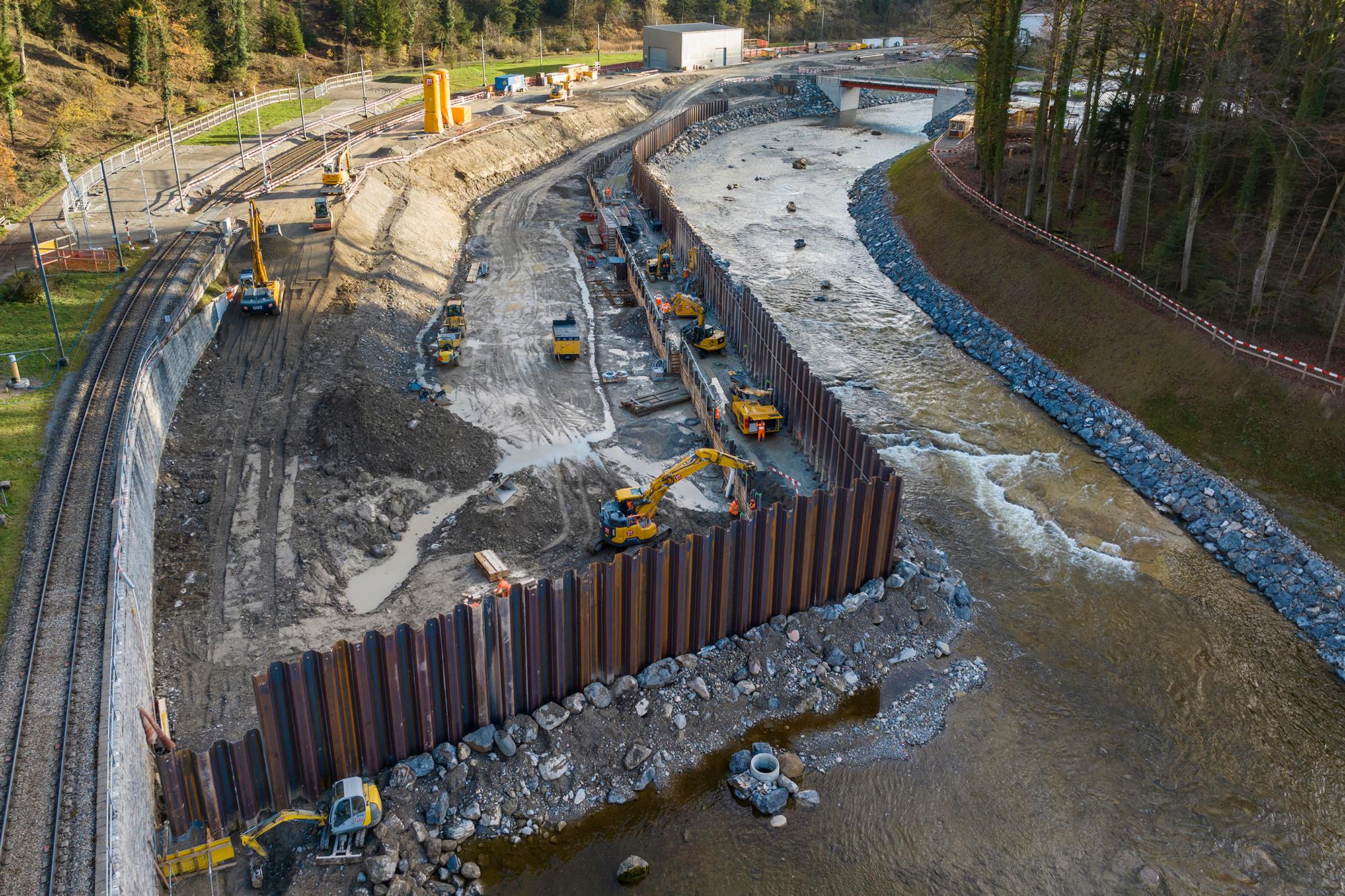 Luftaufnahme der Baugrube, Blick flussaufwärts. Spundwände aus Stahl ragen in der linken Bildhälfte aus einem Damm aus aufgeschüttetem Gesteinsmaterial heraus und bilden eine Reihe bis in den Hintergrund. Links die Bahngleise der SZU, rechts die Sihl.