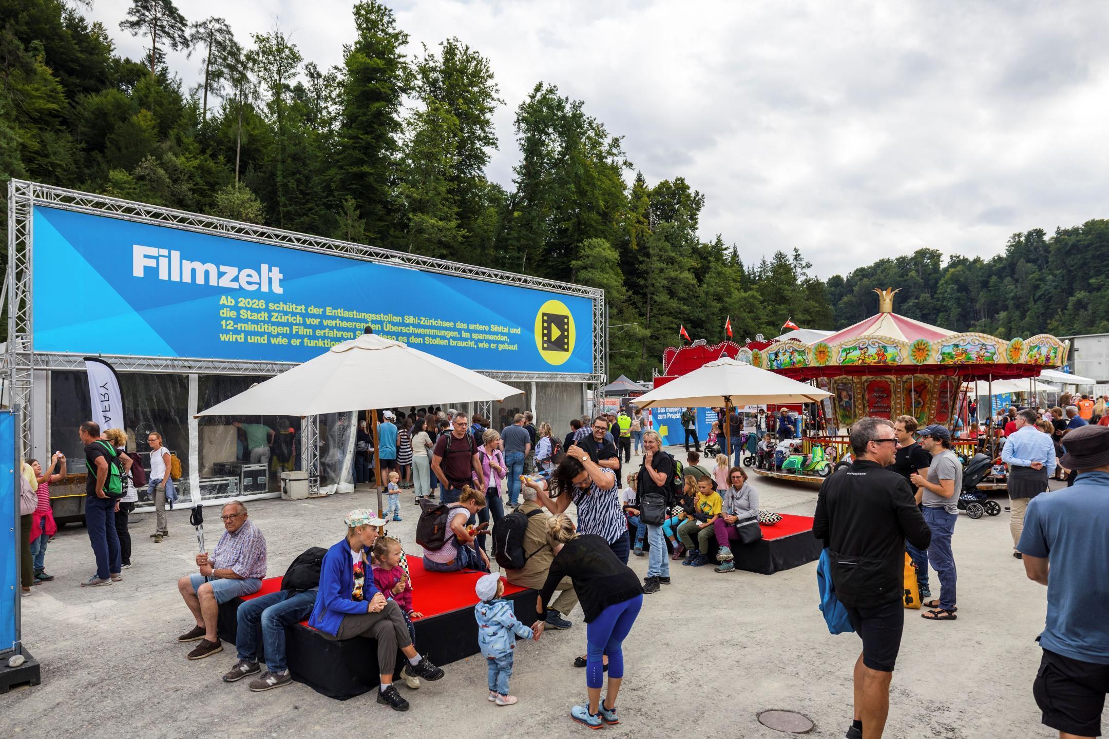 Stehende und sitzende Leute im Vordergrund, hinten das Filmzelt und rechts ein Karussell.