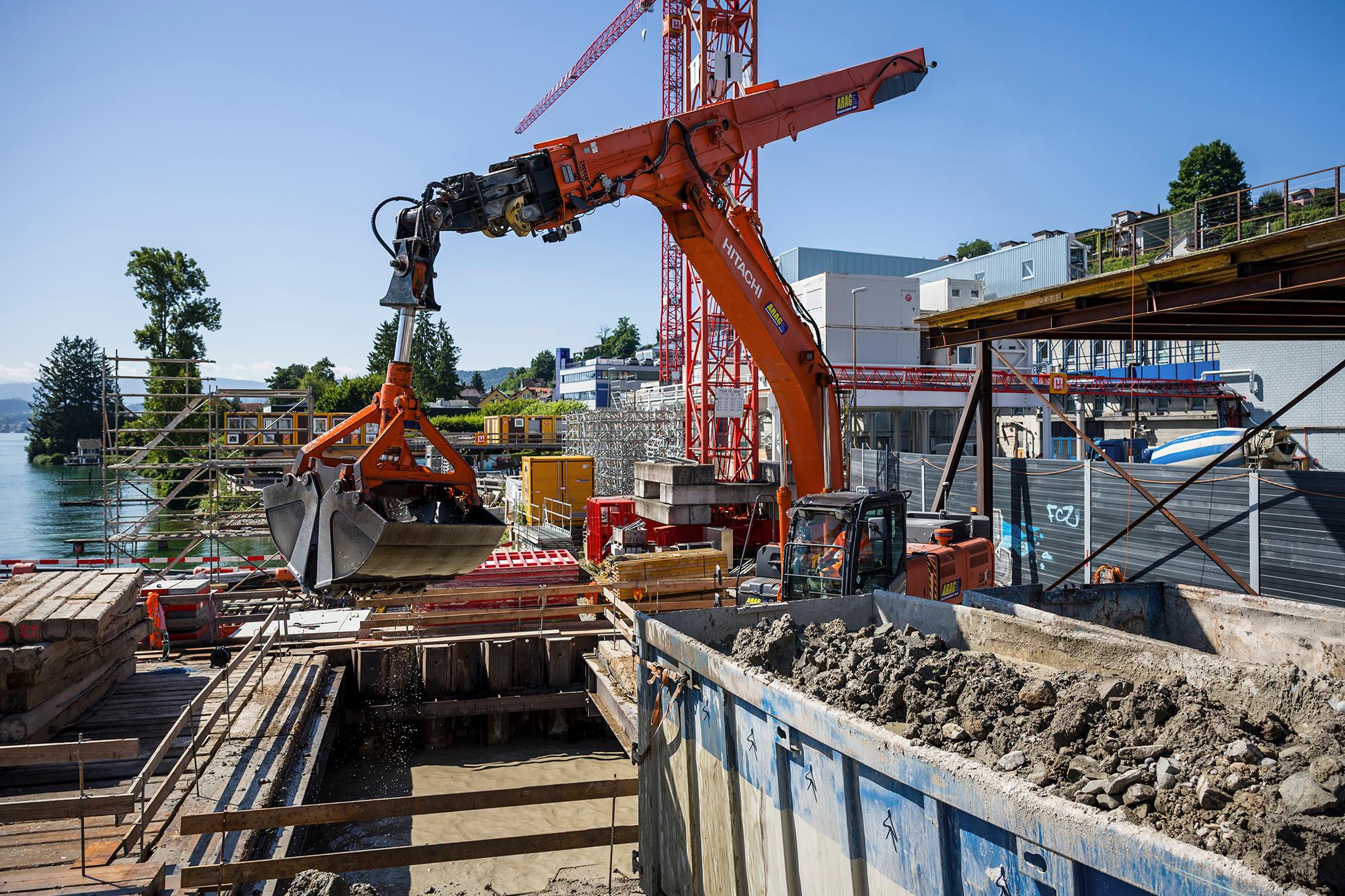 Ein Schalengreifer, der den Seegrund ausbaggert in der Baugrube in der Mitte, oberhalb die Schaufel, aus der das Wasser abtropft und rechts die Mulde mit dem ausgebaggerten Aushubmaterial.