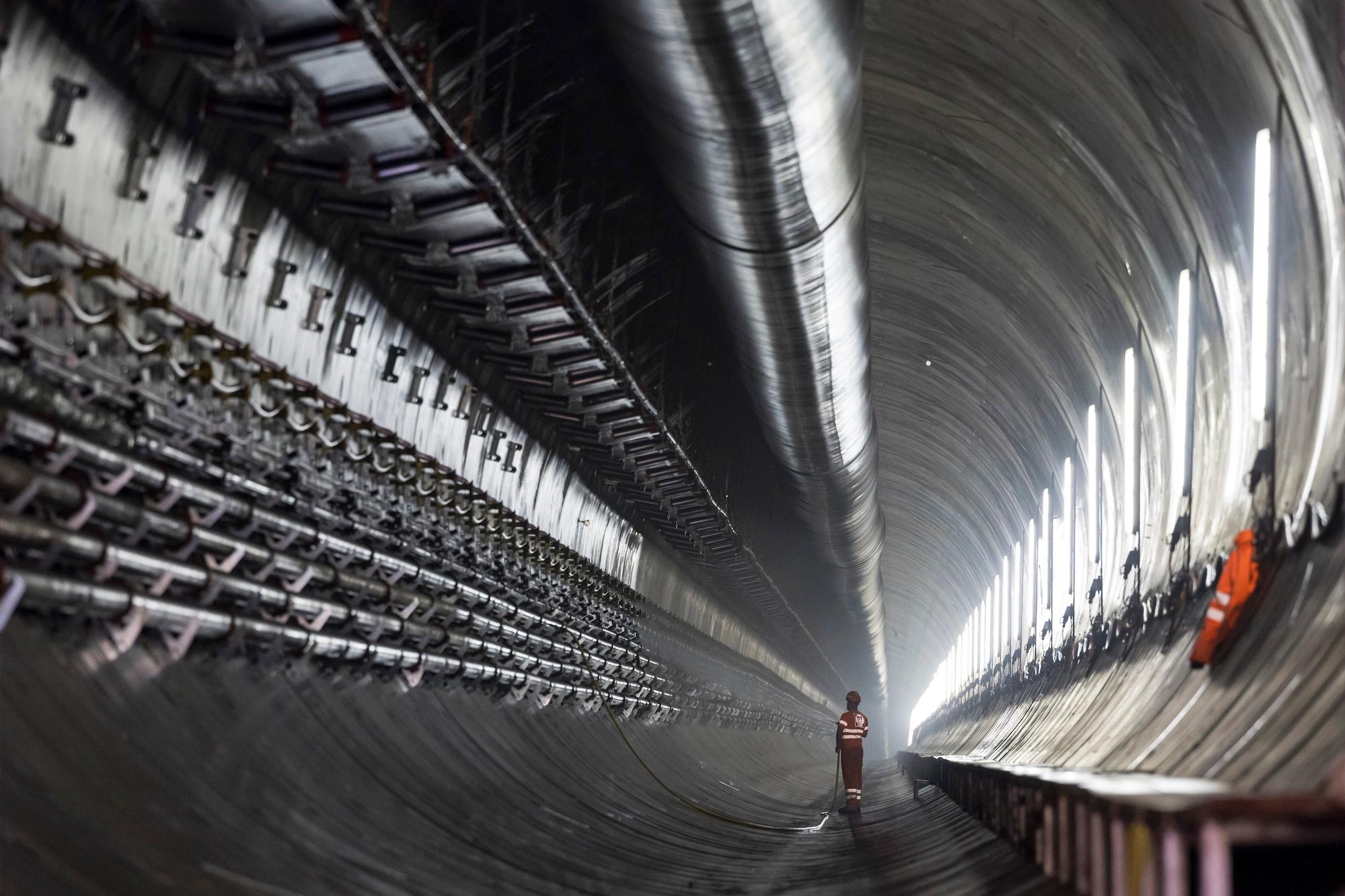 Entlastungsstollen: An den Stolleninnenwänden sind Rohre befestigt und das Förderband, mit dem das Ausbruchmaterial aus dem Stollen gebracht wird. in der Bildmitte steht ein Bauarbeiter mit einem Schlauch.