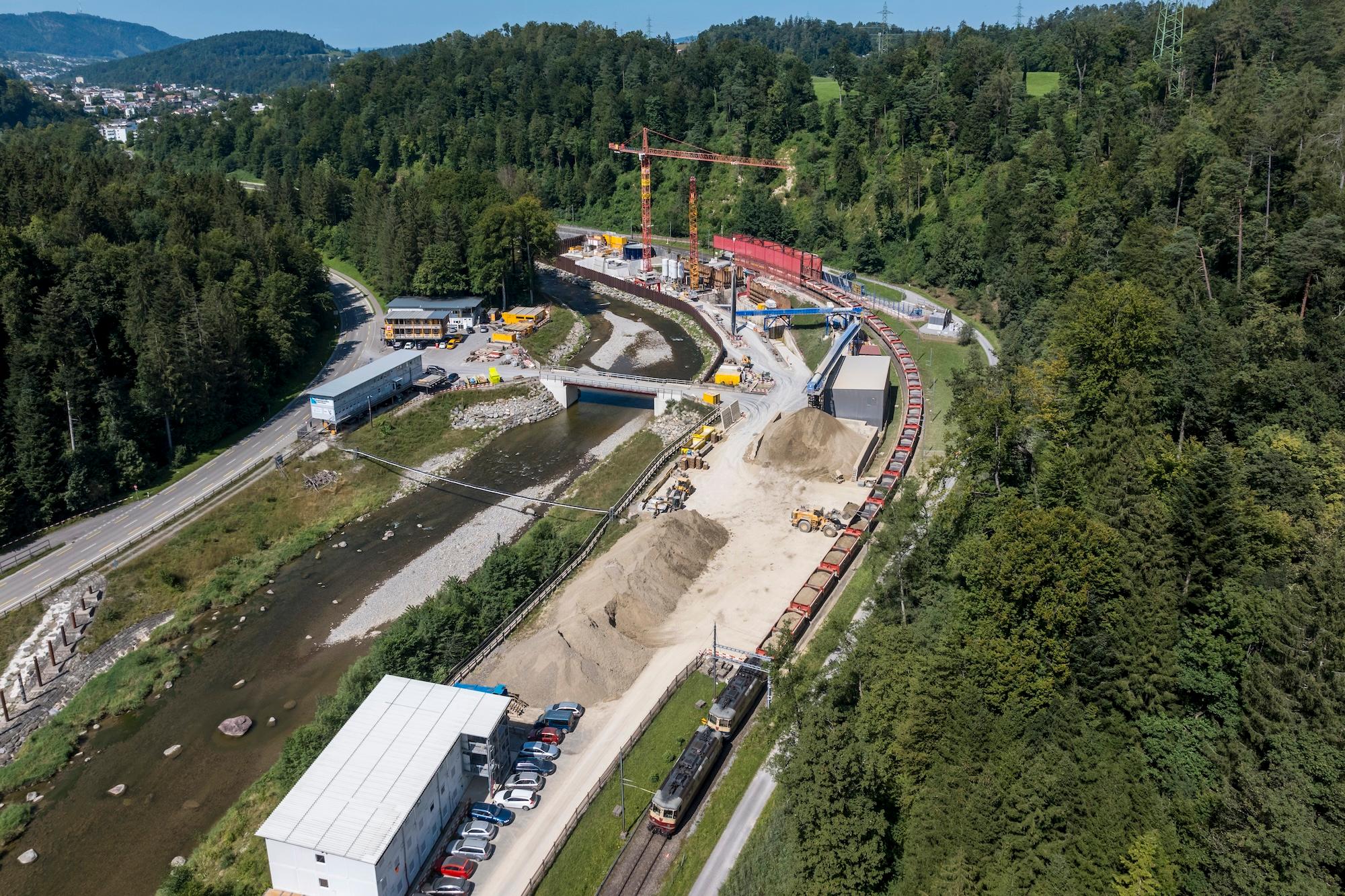 Luftaufnahme der Baustelle des Einlaufbauwerks. Im Hintergrund die Baustelle mit einem Förderband. Dieses transportiert das Ausbruchmaterial aus dem Stollen. Im Vodergrund verlädt ein Bagger das Ausbruchmaterial auf den Zug.
