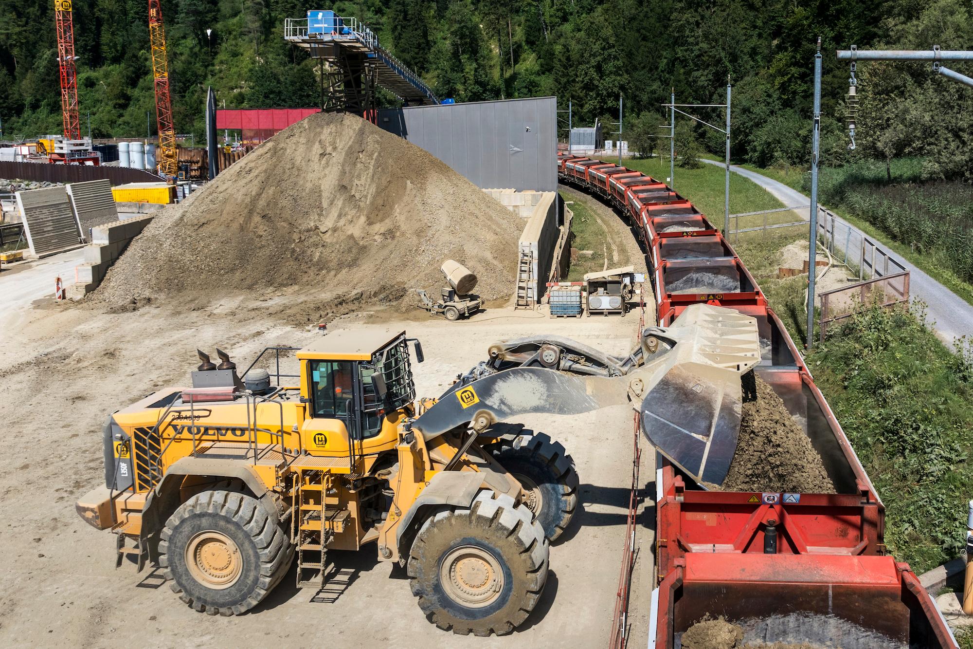 Im Hintergrund des Bildes befindet sich aufgehäuftes Ausbruchmaterial aus dem Entlastungsstollen. Im Vordergrund kippt ein Bagger das Ausbruchmaterial in einen Bahnwagen.