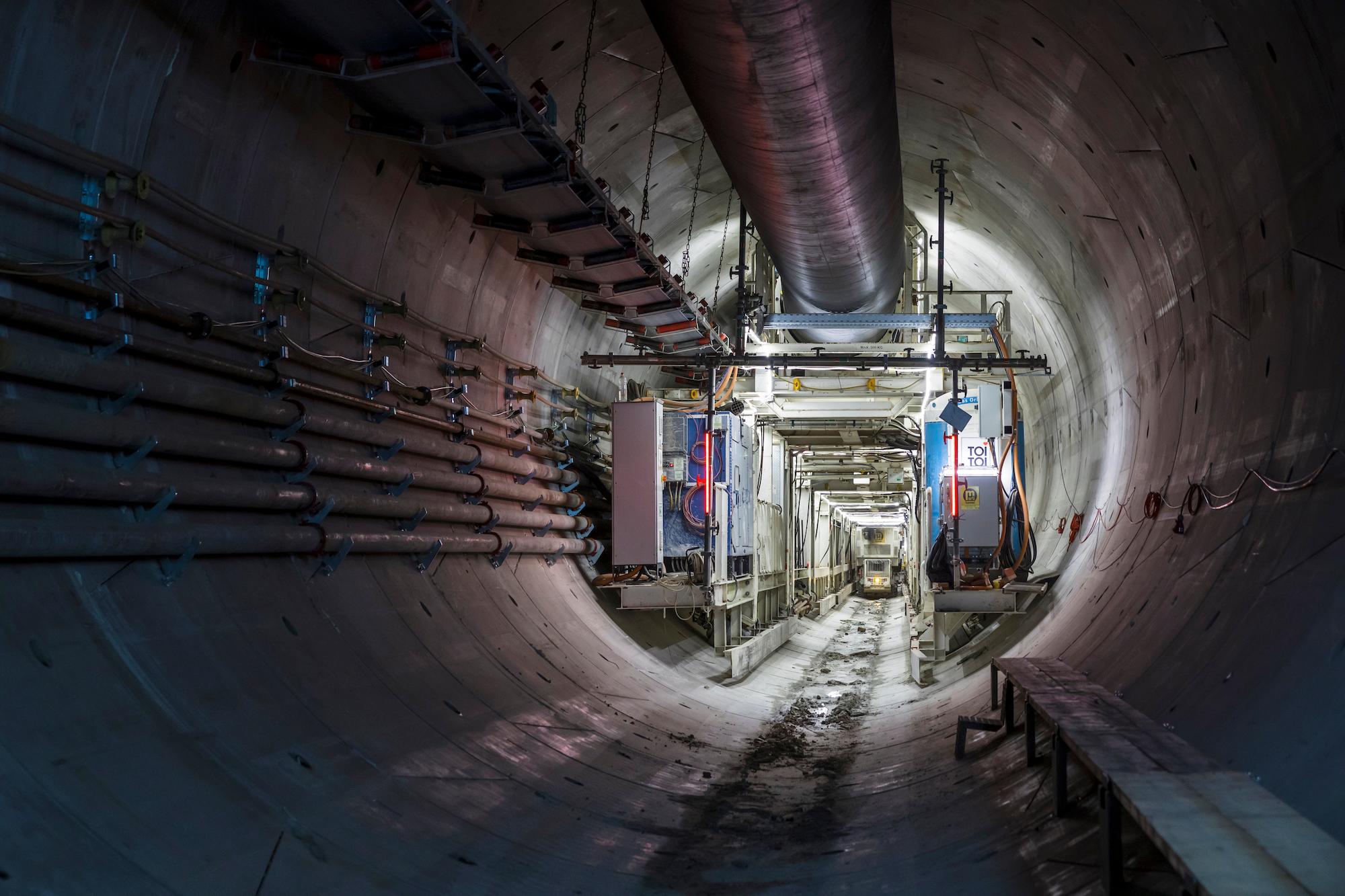 Im Entlastungsstollen sind die Nachläufer der Tunnelbohrmaschine zu sehen. An den Stolleninnenwänden sind Rohre befestigt sowie auch das Förderband, mit dem das Ausbruchmaterial aus dem Stollen hinausbefördert wird.