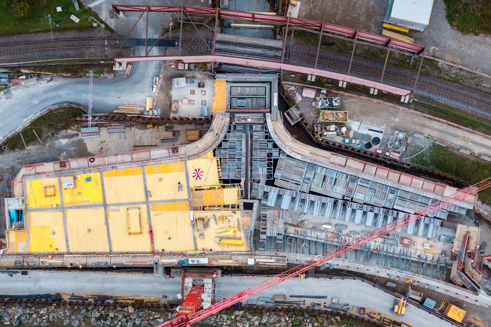 Luftbild des Stollens aus Vogelperspektive, links davon gelbe Schalungsbretter, rechts Baugerüste, gegenüberliegend des Stollens ein roter Baukran