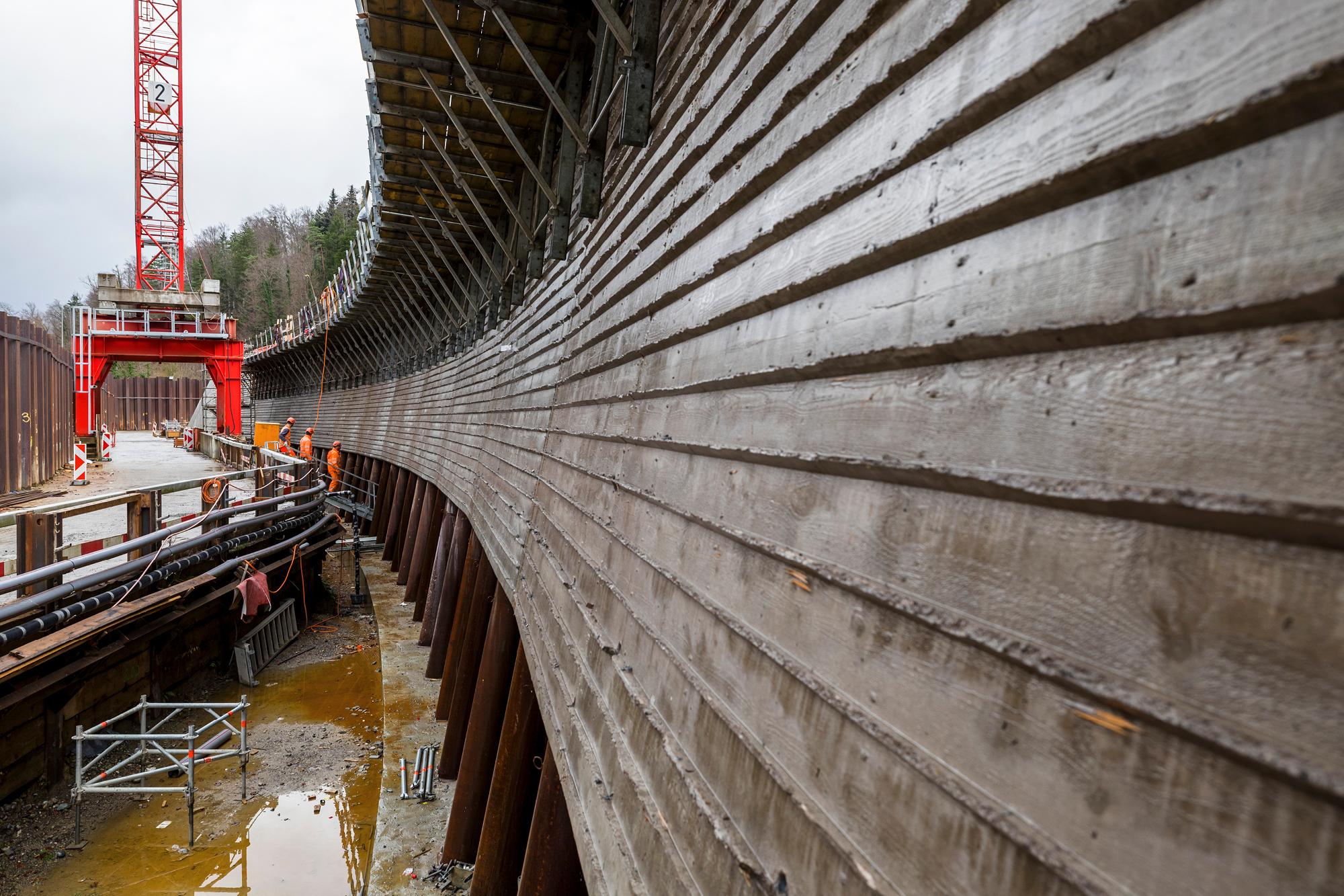 Eine längs gerippte Betonwand, links daneben im Hintergrund ein roter Baukran. und im unteren Bereich der Betonwand die Stahlstützen des Schwemmholzrechens