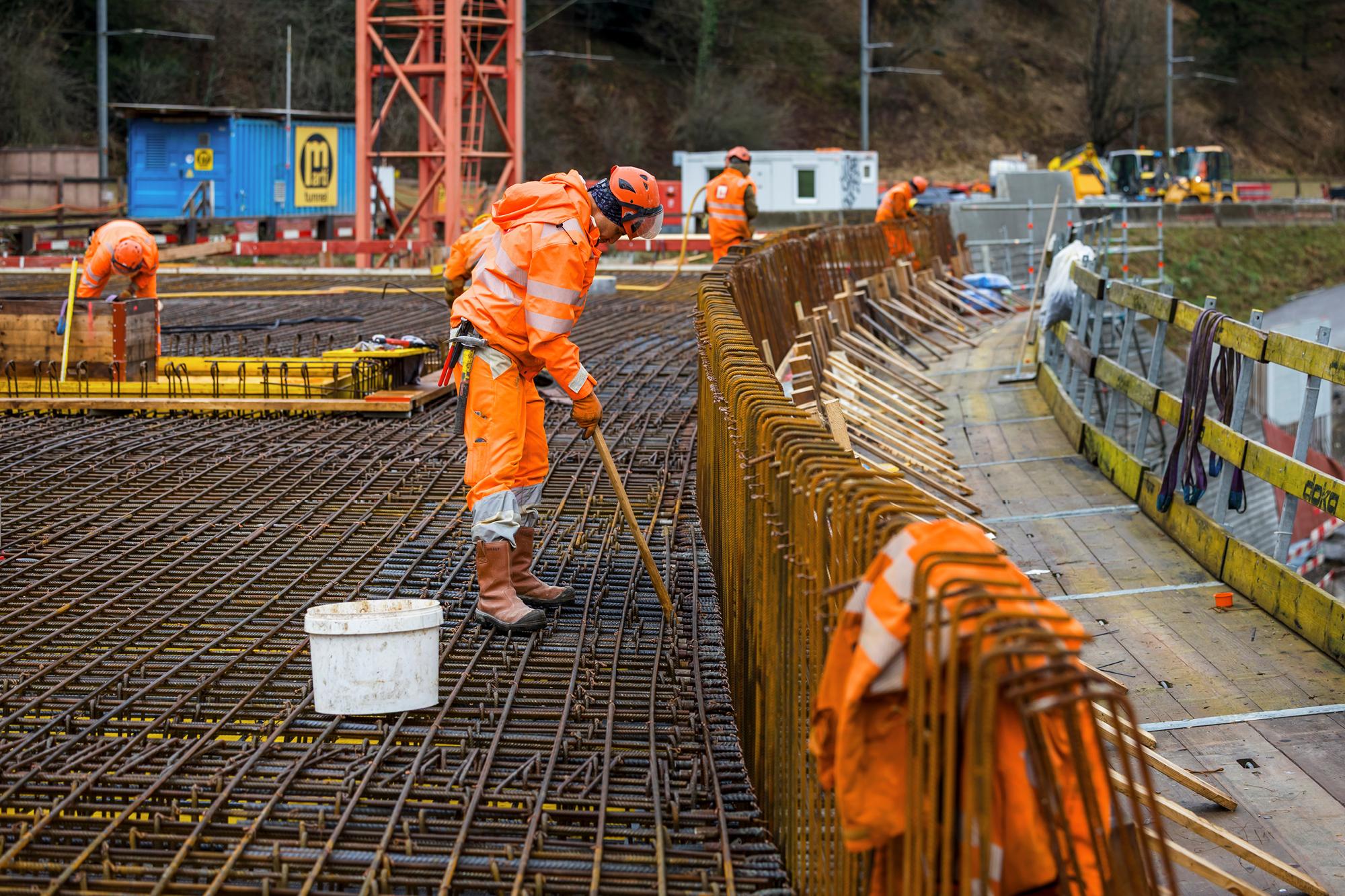 Ein Bauarbeiter steht auf einem Gitter aus rostigen Eisenstangen bei der Verlegung von Armierungseisen. Im Hintergrund weitere Arbeiter und Baucontainer.