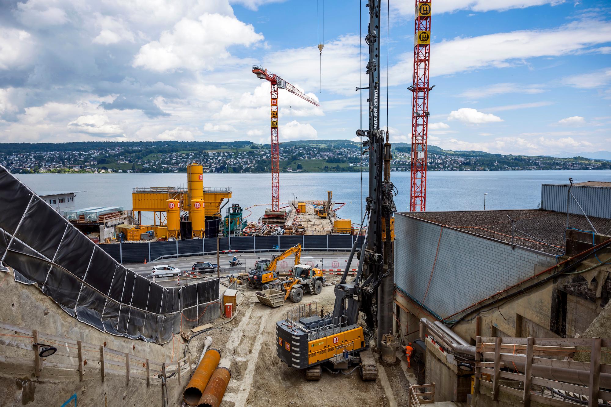 Auf einer Baustelle drückt eine Maschine ein Bohrrohr in den Boden. Im Hintergrund befindet sich der Zürichsee und das gegenüberliegende Seeufer.