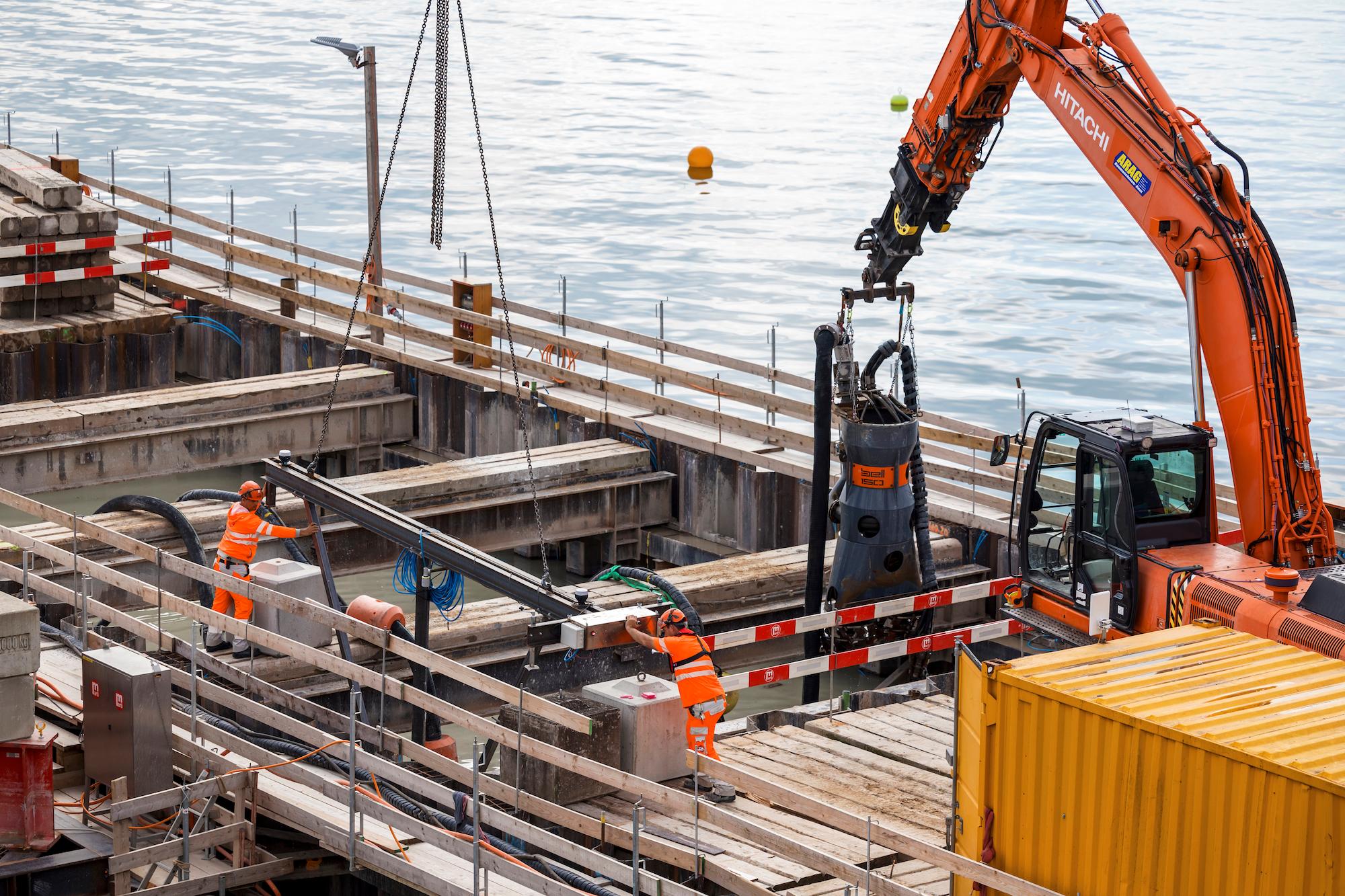 Auf einer Baustelle auf dem Zürichsee hängt ein Stahlelement an Anschlagketten. Zwei Personen mit Helmen und oranger Sicherheitskleidung halten das Stahlelement fest. Rechts davon steht ein Bagger, an dem eine Pumpe befestigt ist.