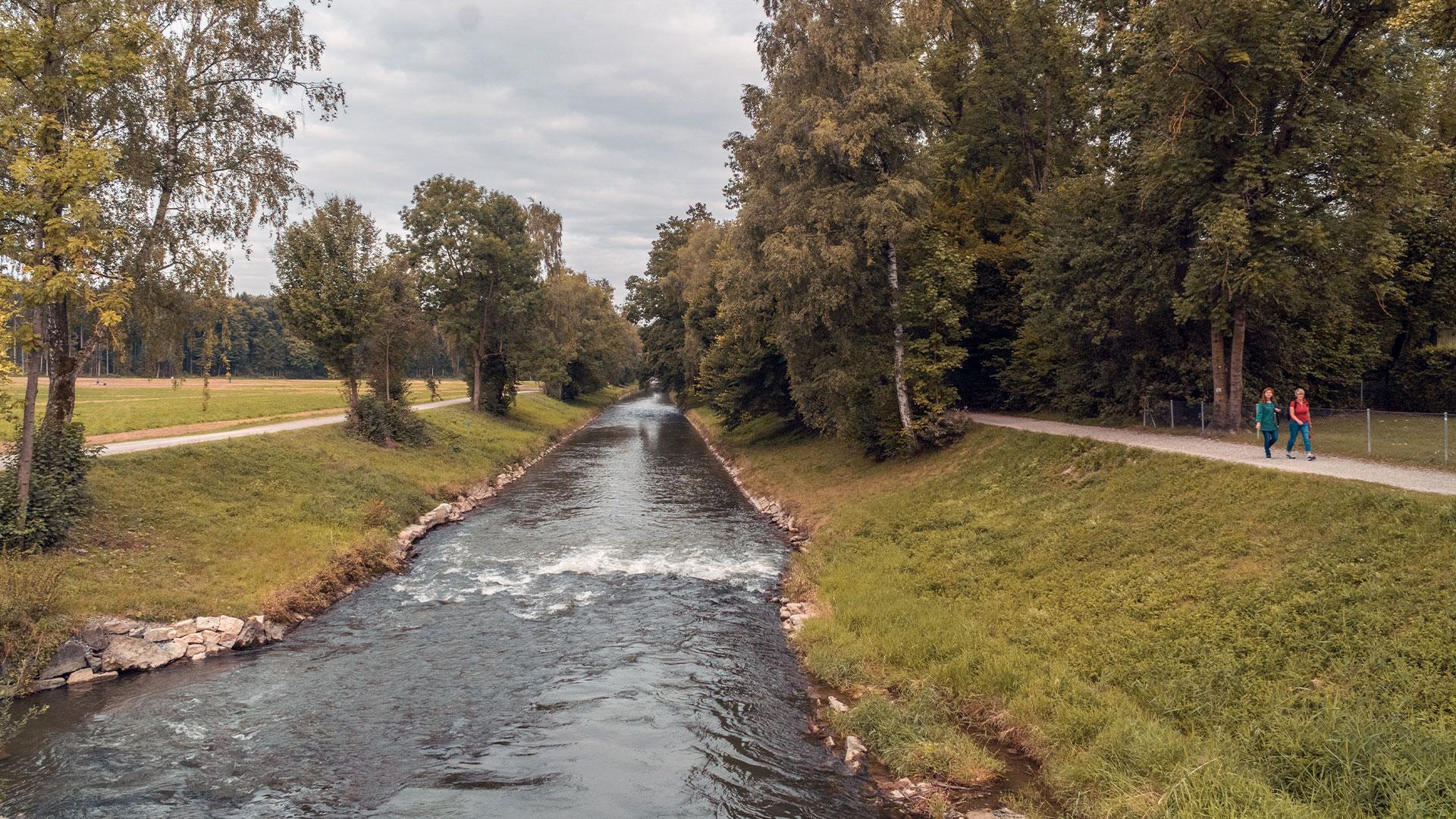 Aufnahme eines Flusses mit deutlich sichtbarer Strömung, gesäumt von breiten Uferwegen und einer Allee, die Raum für Erholung und Spaziergänge bietet.