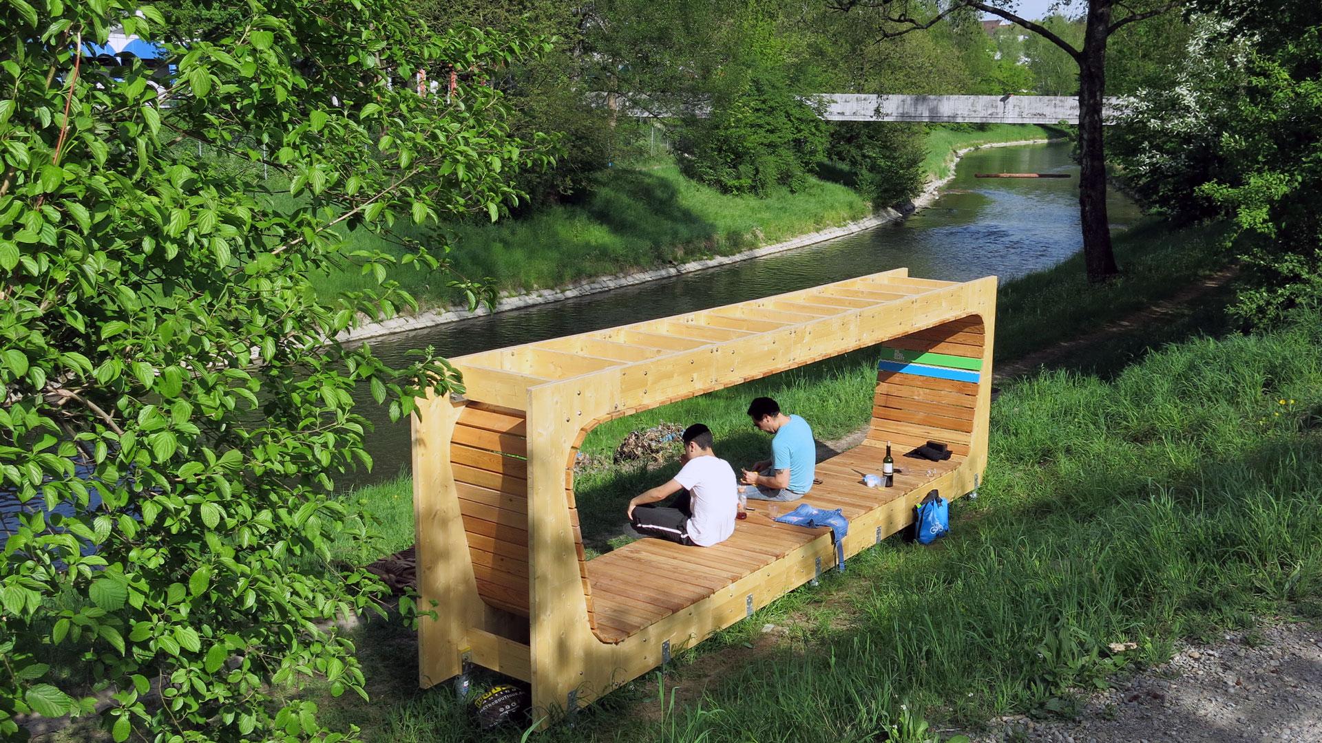 Zwei junge Männer entspannen sich auf einer modernen, hölzernen Sitzskulptur am Ufer eines Flusses, umgeben von grüner Wiese und Blick auf den Flusslauf.
