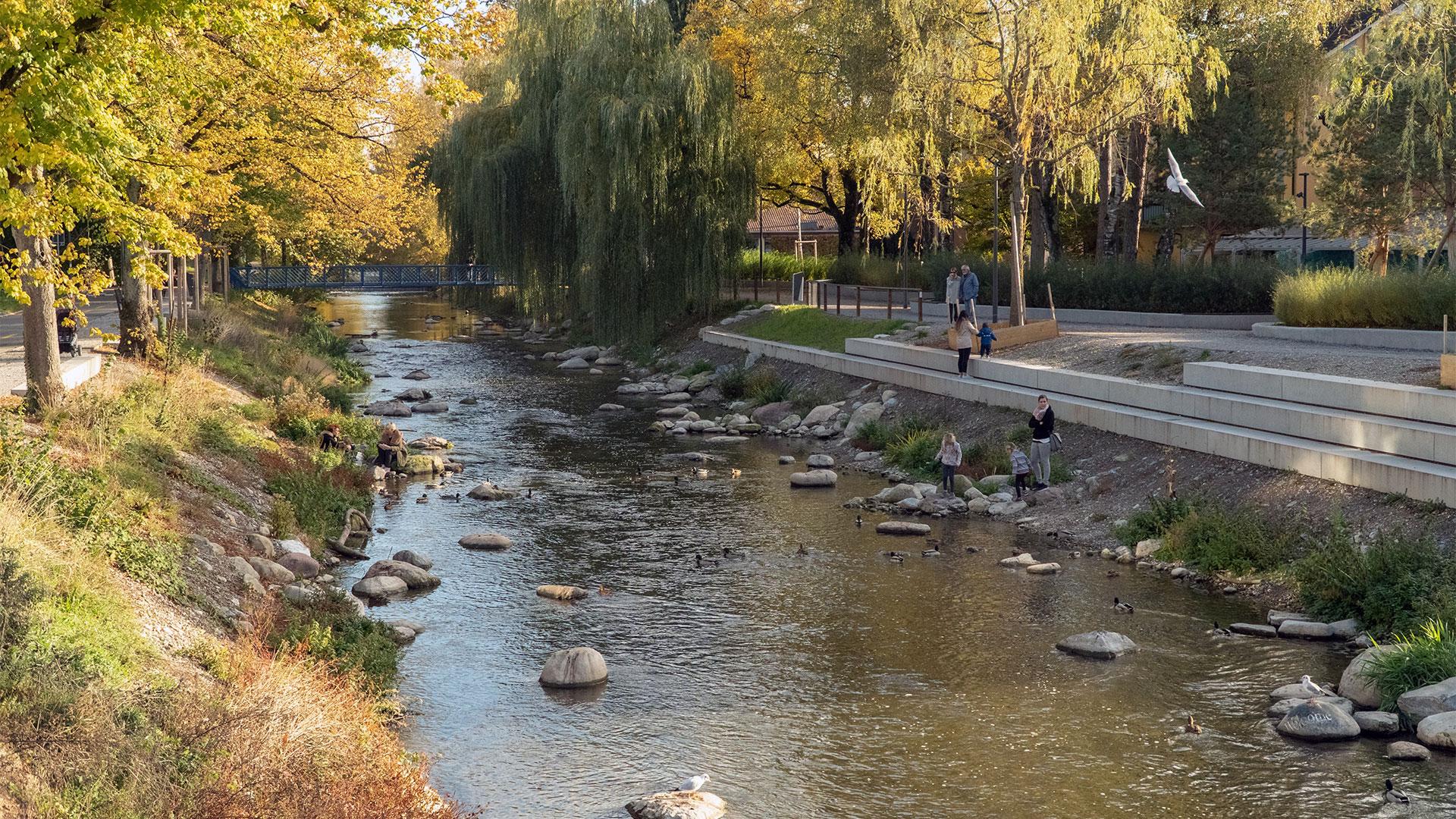 Neu gestaltetes Ufer eines Flusses, mit Betonterrasse und Treppenstufen, die zum flachen Wasser mit Steinen führen; mehrere Familien halten sich am Ufer auf.