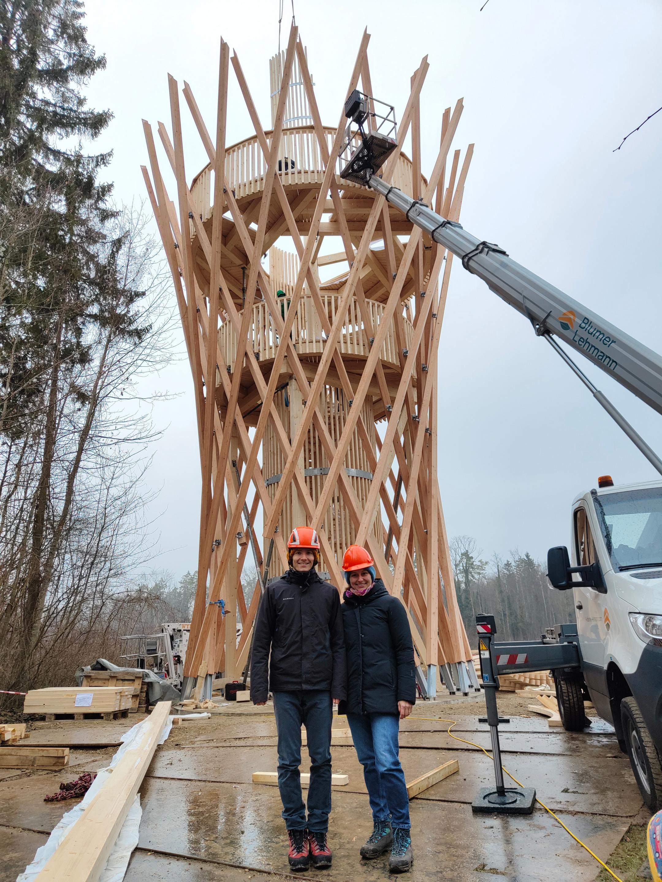 Das Bild zeigt die Baustelle des fast fertig gebauten Holzturms. Rechts im Bild ist ein Fahrzeug mit Hebebühne zu sehen. Die beiden ehemaligen Studierenden stehen im Vordergrund, beide mir orangem Bauhelm, und blicken freudig in die Kamera.