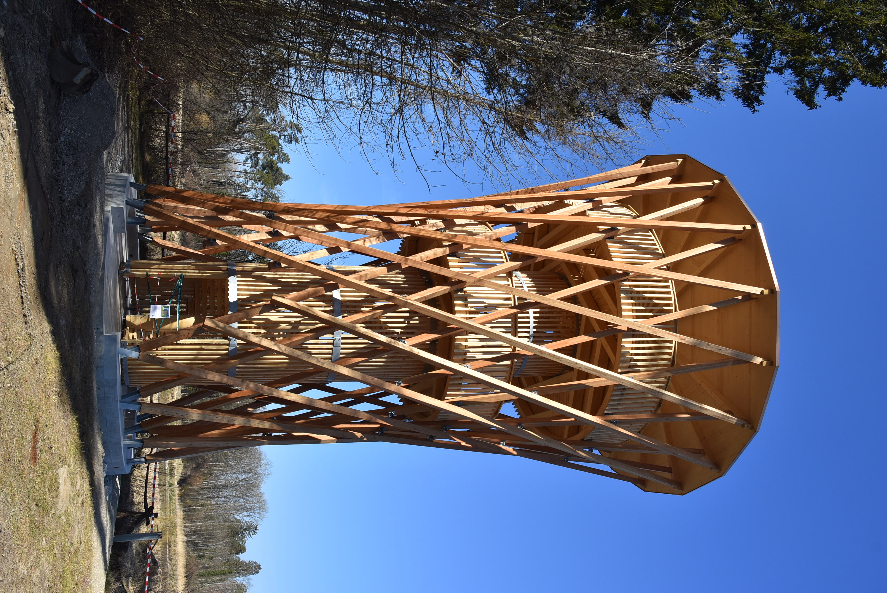 Aussenansicht des Turms vom Zugangsweg her. Das Innere des Turms mit Wendeltreppe und Plattformen ist mit geraden, senkrechten Holzlatten verkleidet. Die Aussenhülle wird durch eine hyperbolische Gitterstruktur aus langen Lärchenhölzern gebildet.