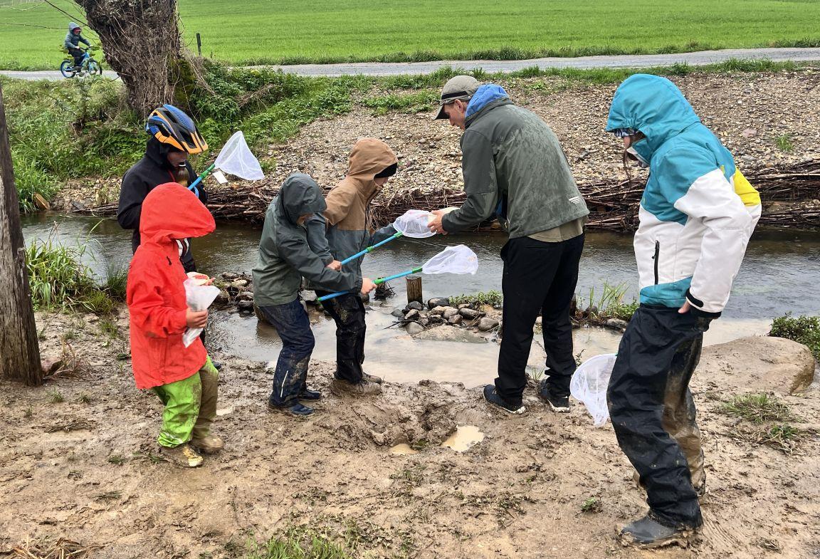 Vier Kinder in Regenkleidern stehen am Bachufer und halten kleine Netze an langen Stäben in der Hand. Ein Mann erklärt einem der Kinder, was sich im Netz befindet.