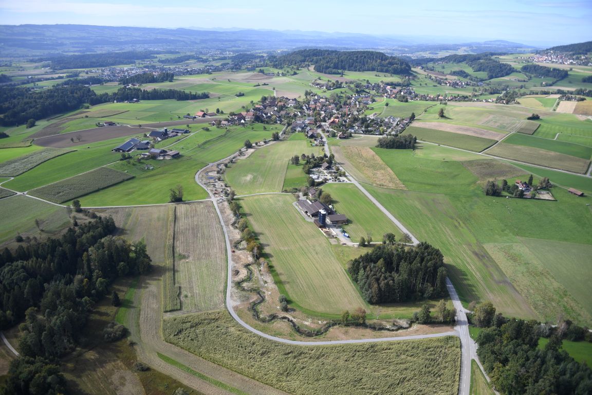 Eine Aufnahme aus der Luft zeigt einen Bach, der in Schlaufen durch die Landschaft mäandriert. In der Umgebung sind Felder, Wälder, Strassen und Siedlungen zu sehen.