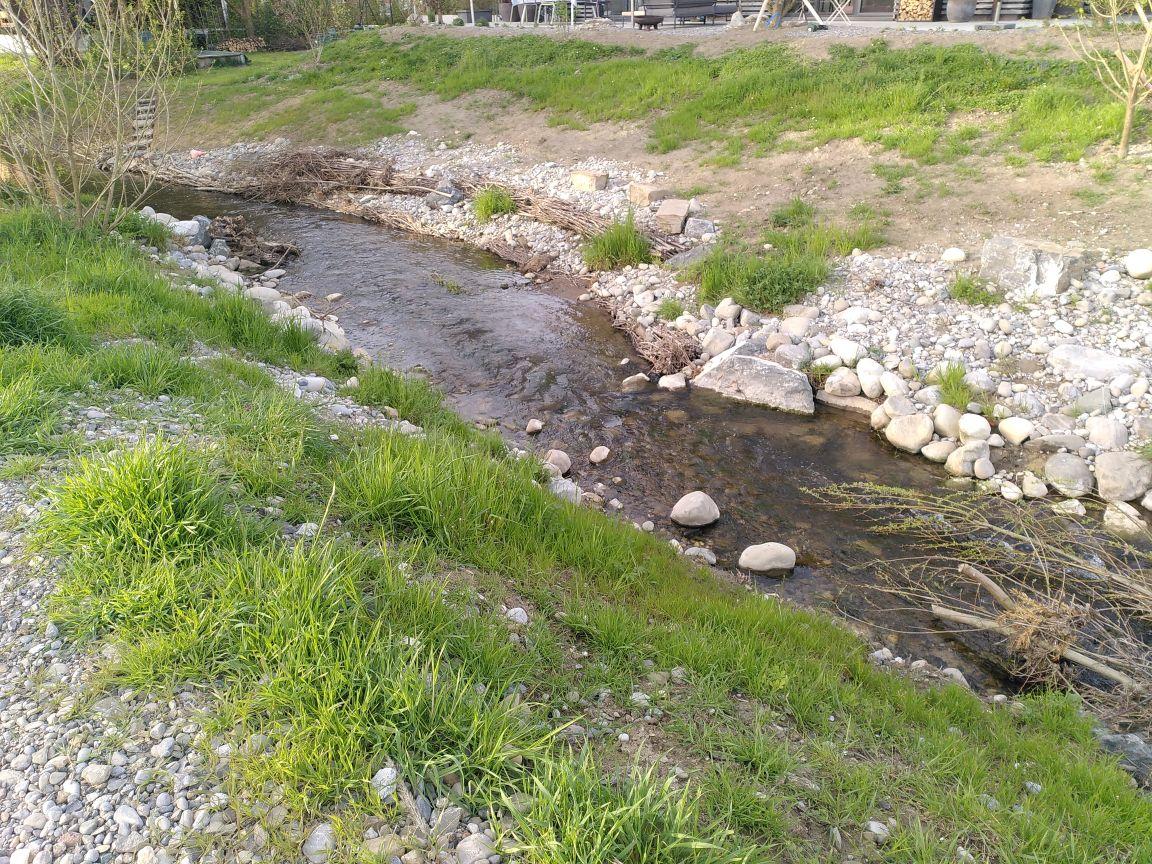 Das Foto zeigt einen Bach mit Kies, Steinen und Astbündeln entlang der Ufer und im Wasser.