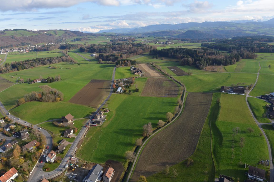 Eine Aufnahme aus der Luft zeigt einen Bach, der begradigt durch die Landschaft fliesst. In der Umgebung sind Felder, Wälder, Strassen und Siedlungen zu sehen.