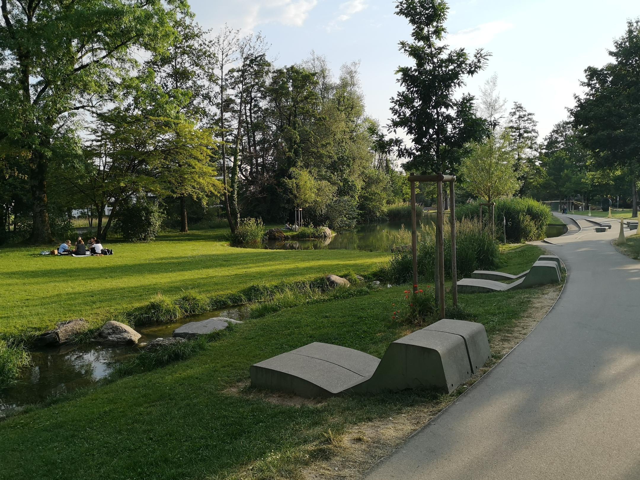 Neben dem Spazierweg durch den Stadtpark Uster fliesst die Aa. Moderne Betonelemente in Liegestuhlform bieten Sitzmöglichkeiten auf der grünen Wiese.
