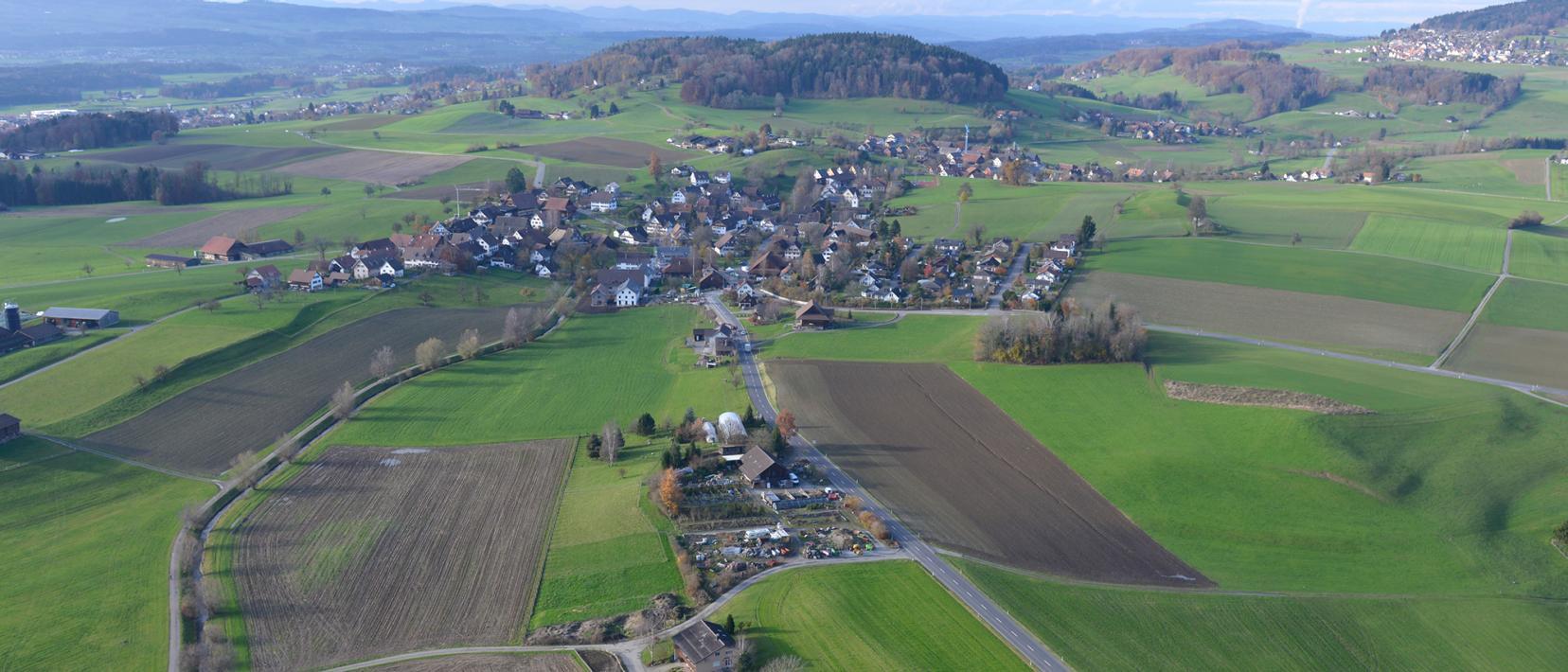 Luftaufnahme des Dorfes Rifferswil, das von Feldern und Äcker umgeben ist. Die stark kanalisierte Jonen verläuft durch das Dorf. 