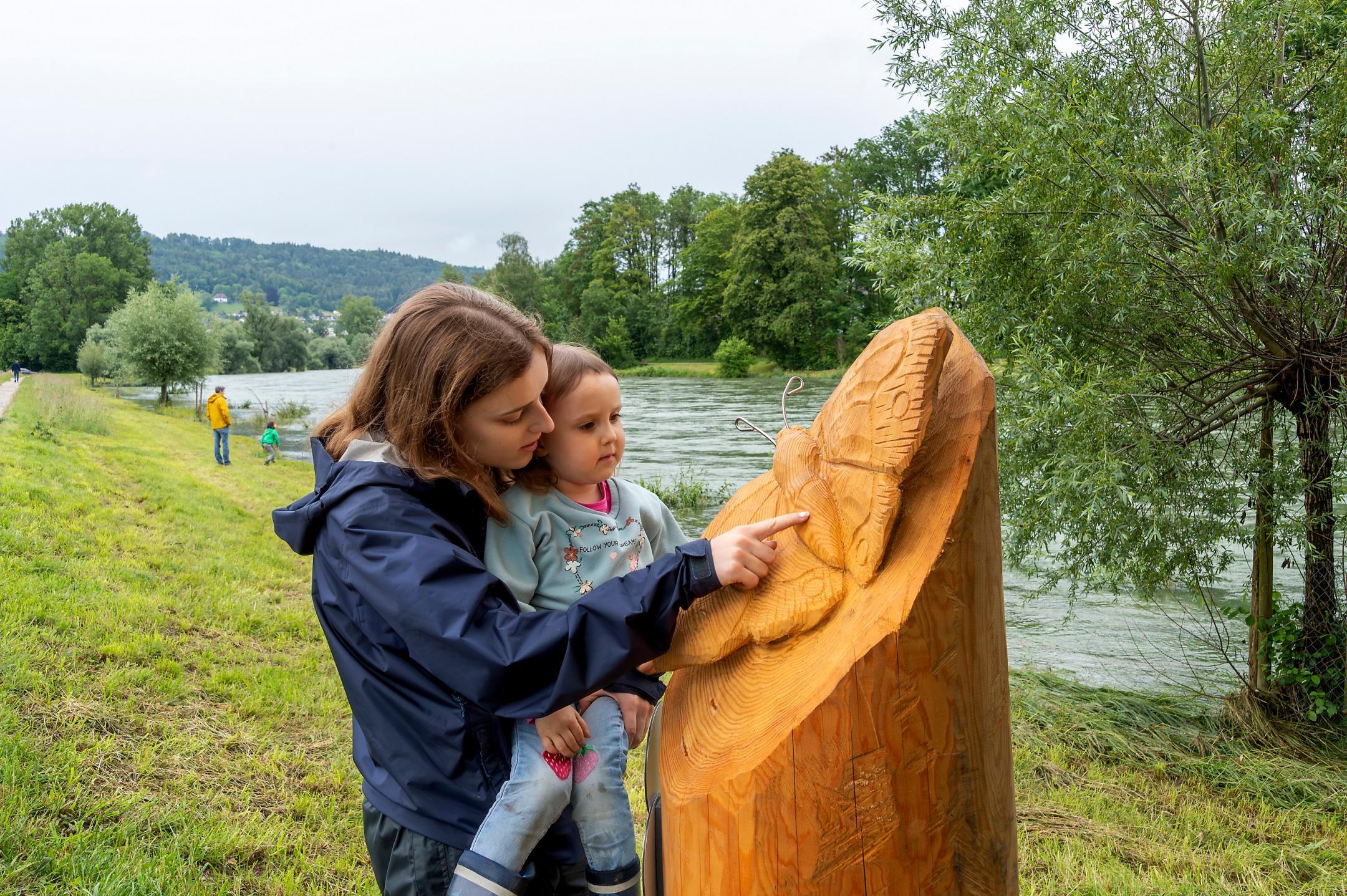 Eine Frau mit einem Kind auf dem Arm bewundert eine geschnitzte Schmetterlingsfigur an der Limmat.