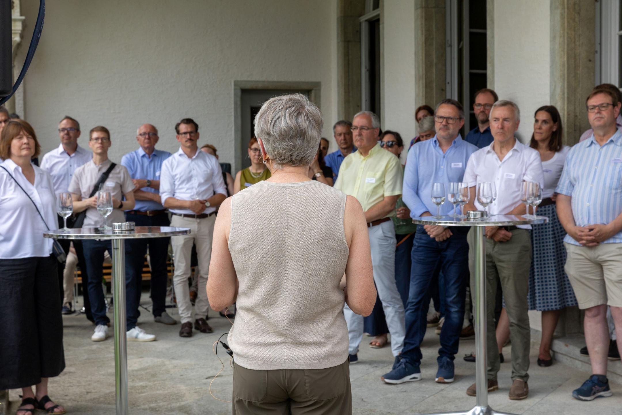 Eine Frau mit dem Rücken zur Kamera spricht vor einer Gruppe von Gästen