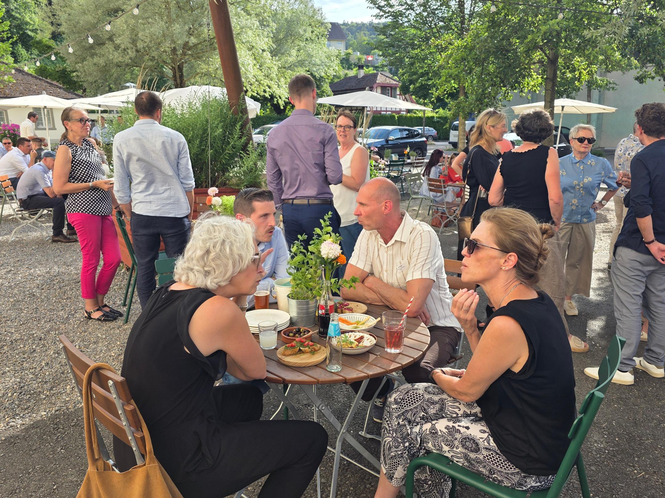 Im Vordergrund sitzen vier Menschen an einem Tisch und unterhalten sich, im Hintergrund ein Platz, auf dem sich weitere Menschen unterhalten.