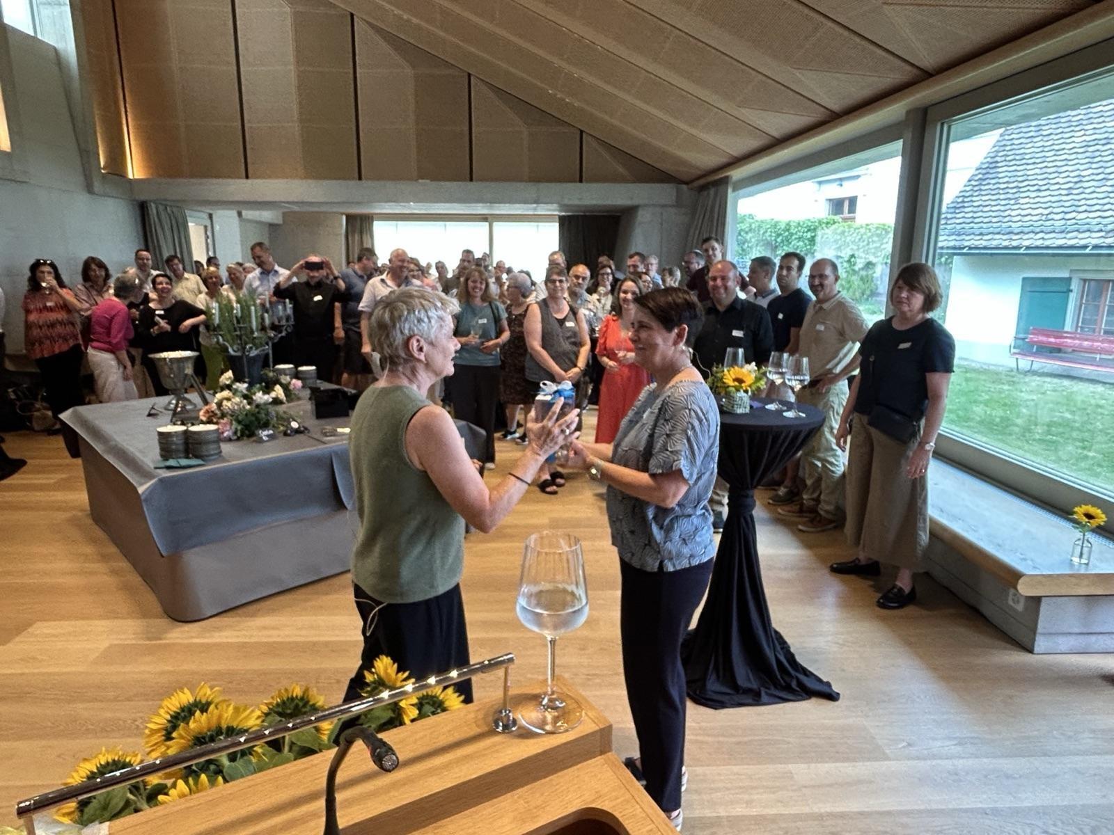 Zwei Frauen stehen in einem Saal vor einer Gruppe von rund 80 Menschen. Die eine Frau übergibt der anderen ein Geschenk.