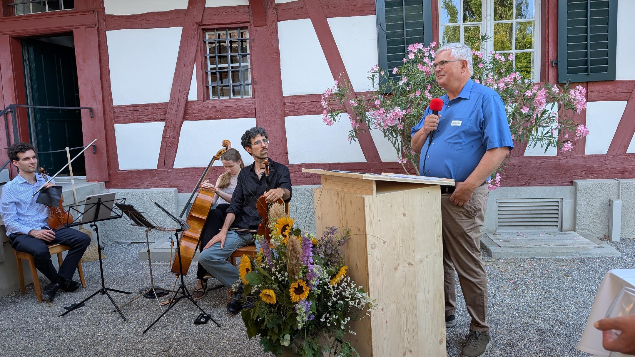 Link ein Mann an einem Rednerpult mit Mikrofon, rechts neben ihm sitzt ein Streicher-Trio