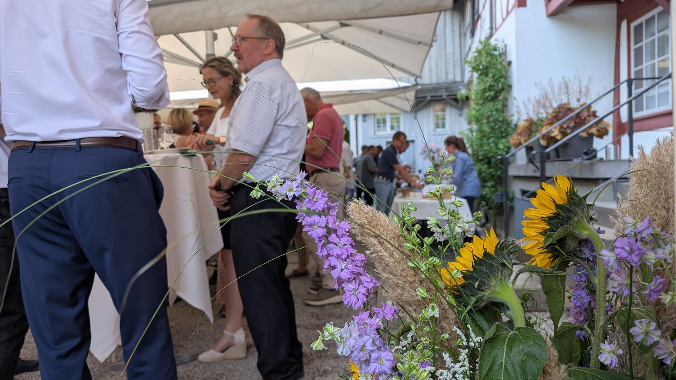 Blick auf eine Gruppe Menschen in einem Hof beim Apero, im Vordergrund Blumen